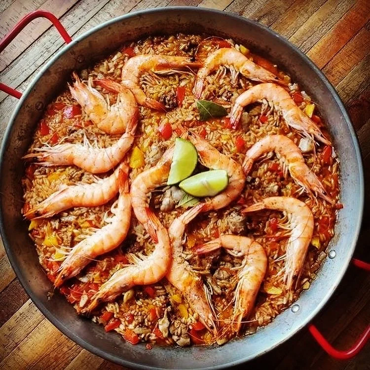 Shrimp and rice dish with chopped vegetables in a black skillet garnished with lime wedges and a bay leaf.