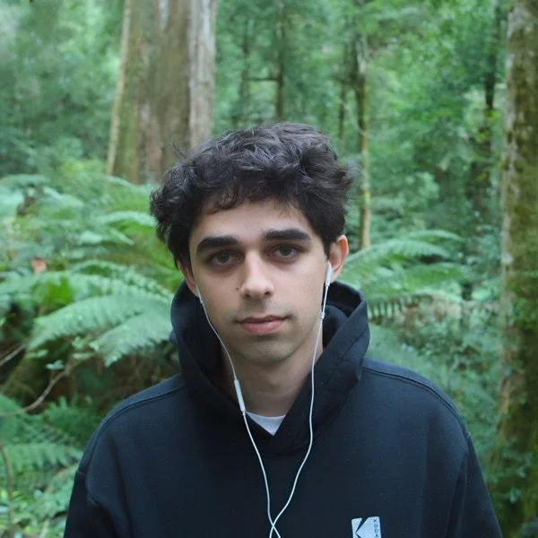 Young man with dark curly hair and light skin wearing a black hoodie and white earphones, standing in a lush green forest.