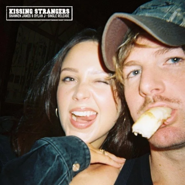 Two young adults, a woman with dark hair and a man with blond hair and a cap, are taking a close-up selfie. The woman is winking and sticking out her tongue, while the man is biting into a banana. There is a small text overlay in the top left corner that reads 'Kissing Strangers' and mentions a single release by Shannen James & Dylan J.