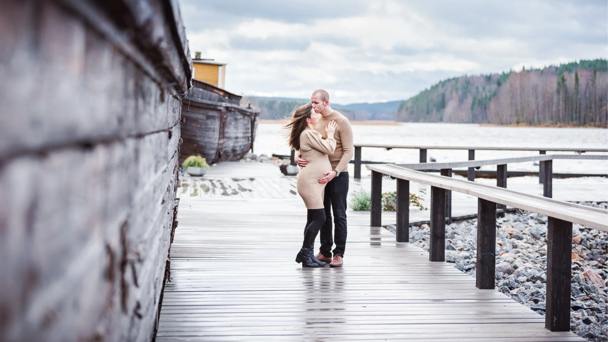 Pariskunta seisoo laiturilla pilvisenä päivänä, mies silittää raskaana olevan puolisonsa vatsaa.