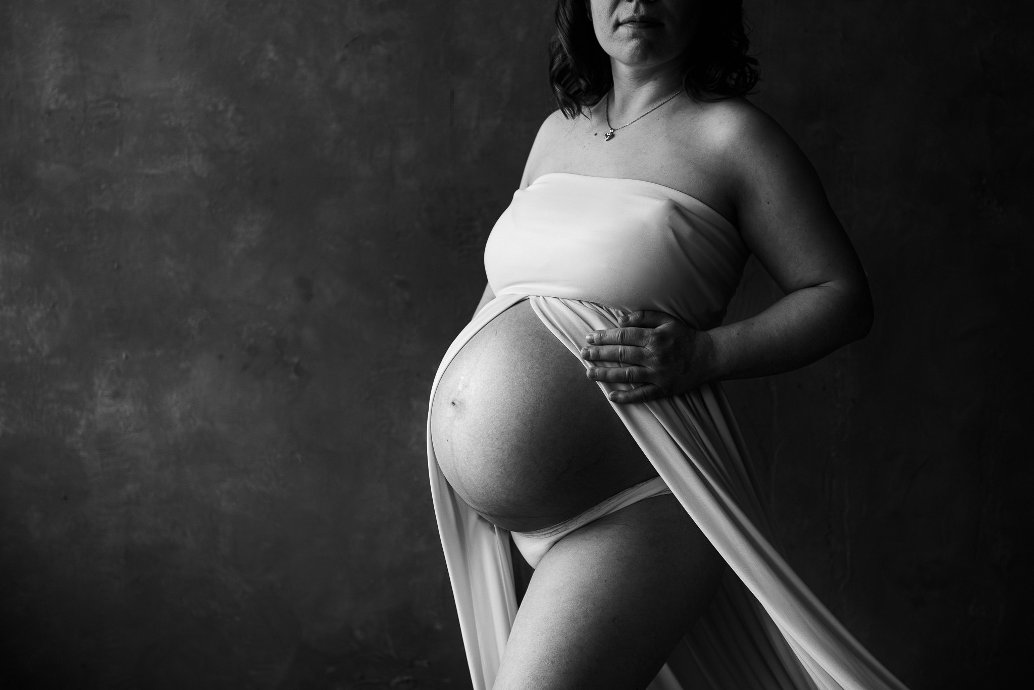 Tummasävyisessä studiossa pehmeä valo piirtää näkyviin naisen raskausvatsan.
