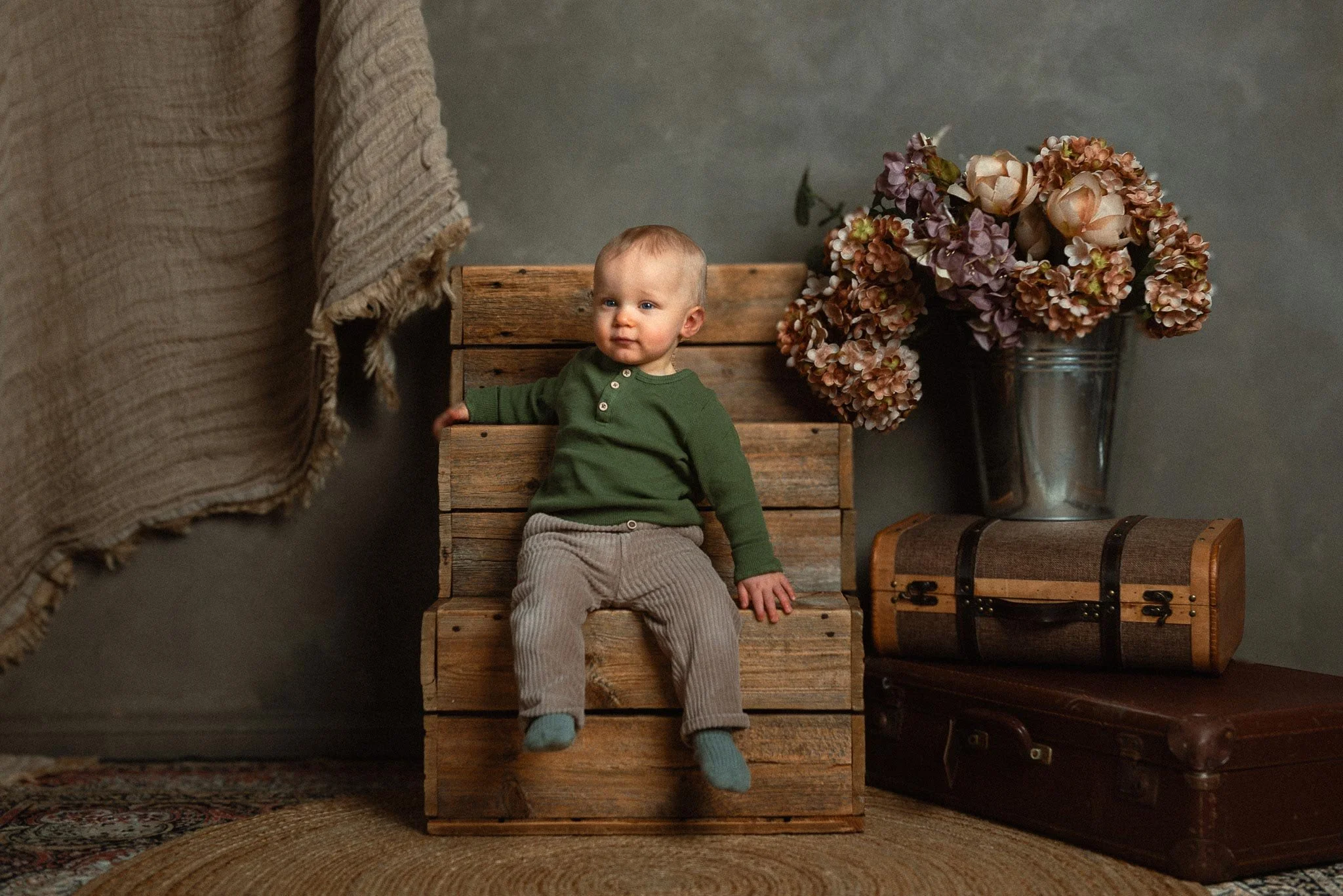 Vihreäpaitainen ja harmaahousuinen taapero istuu puulaatikon päällä, vieressä on kaksi vanhaa matkalaukkua ja niiden päällä sangossa iso kimppu kukkia.