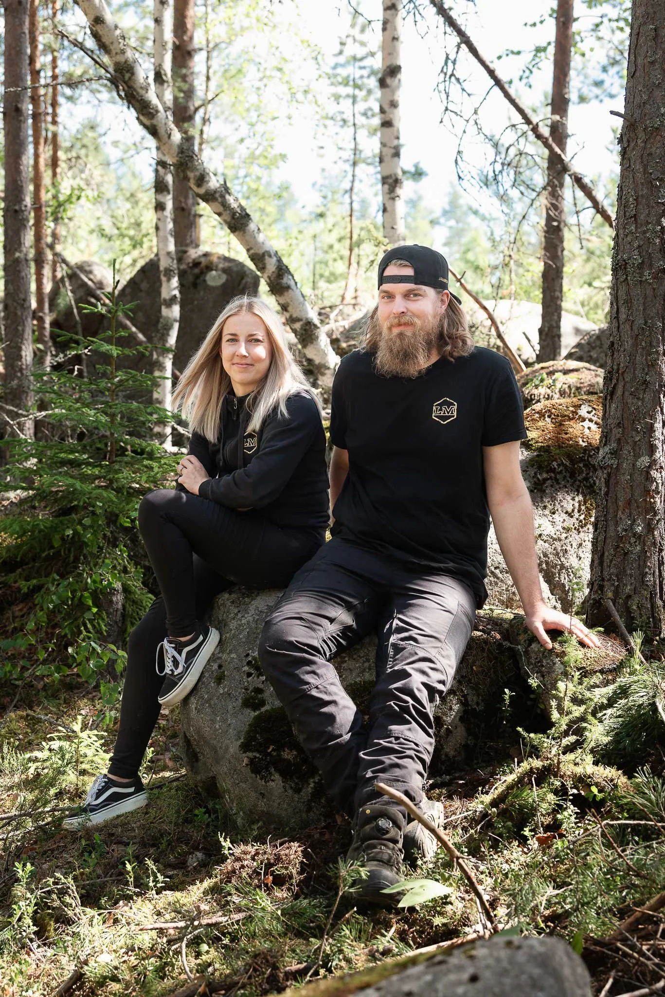 Mustiin LM-logoilla varustettuihin työvaatteisiin pukeutuneet vaaleahiuksinen nainen ja parrakas lippalakkipäinen mies istuvat kivellä metsässä aurinkoisena päivänä.