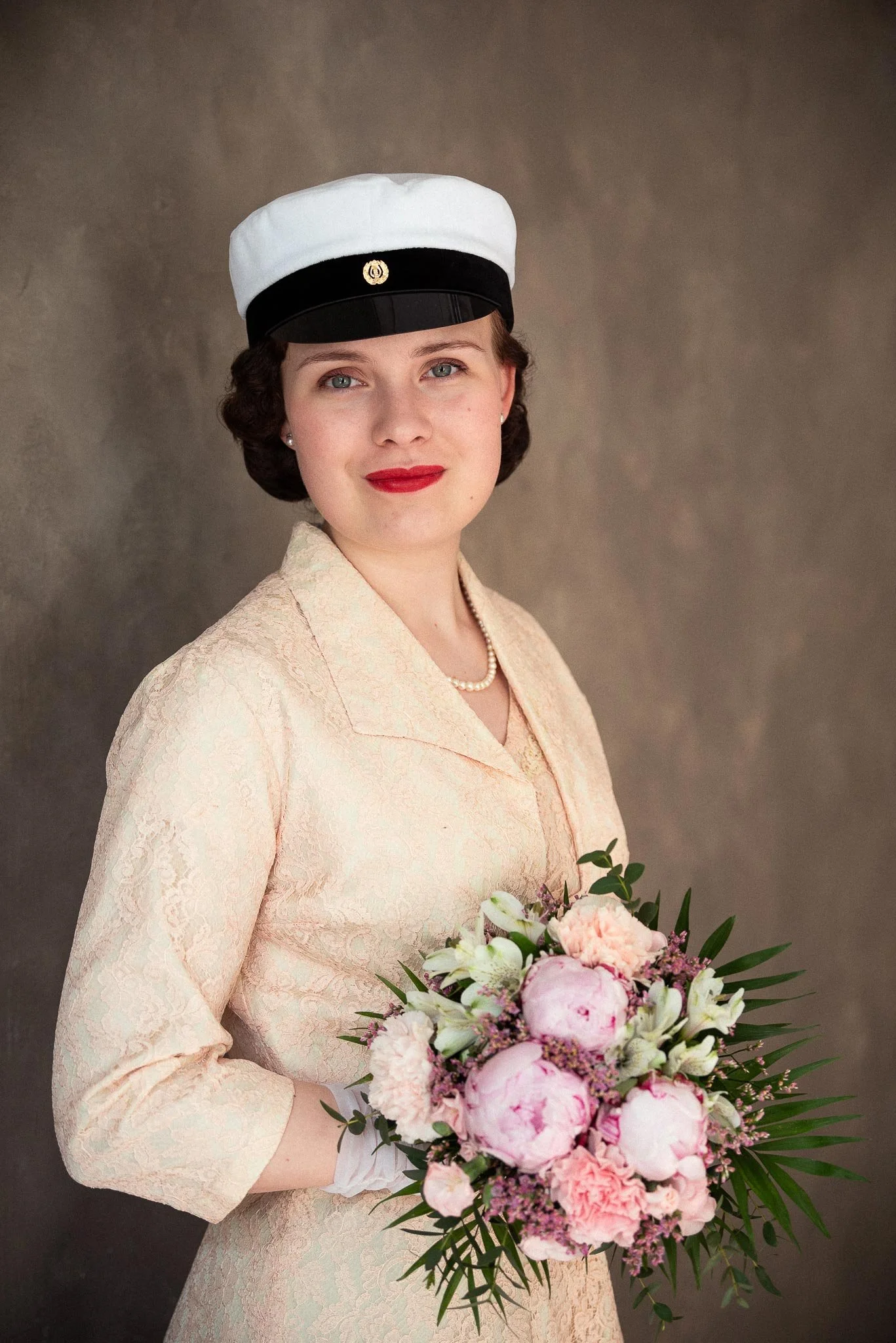 Kermanväriseen kirjailtuun juhlapukuun pukeutunut nuori nainen seisoo kalkkimaalatun ruskean seinän edessä ylioppilaslakki päässään ja kädessään kukkakimppu.