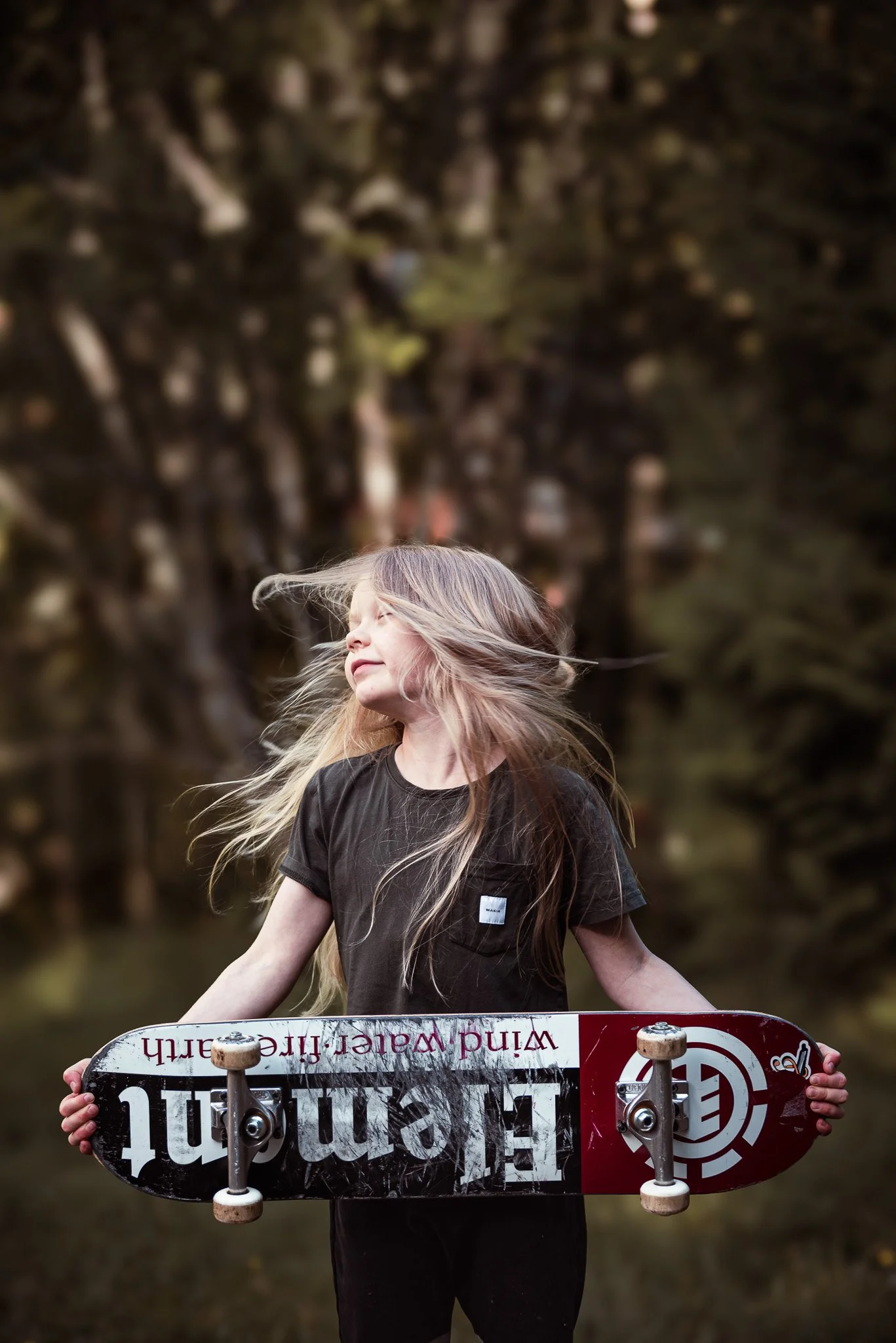 Lapsi seisoo skeittilautaa pidellen ja hulmauttaa pitkiä hiuksiaan.