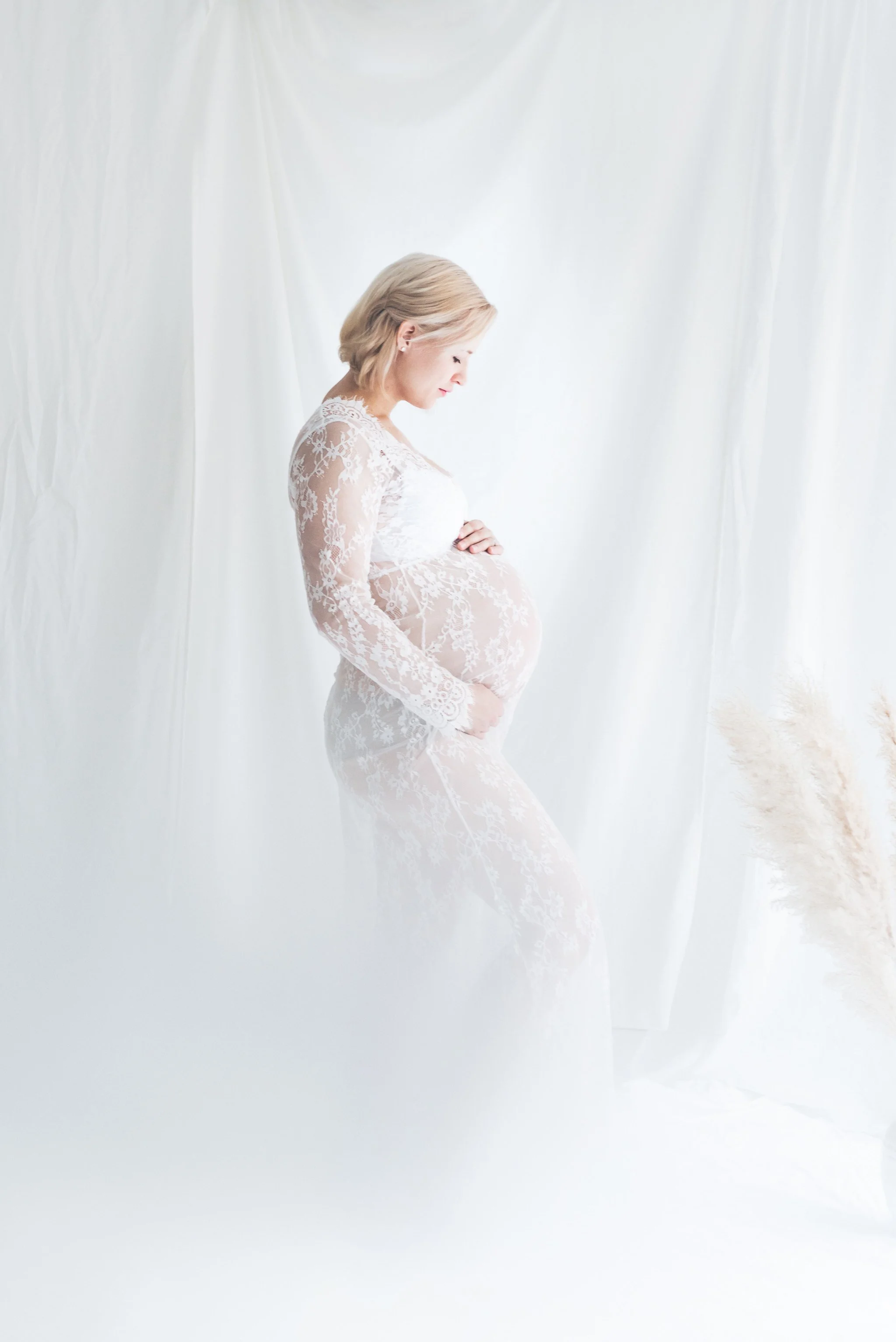 Raskaana oleva nainen seisoo sivuprofiilissa vaaleassa studiossa ja pitelee vatsaansa yllään valkoinen pitsimekko.