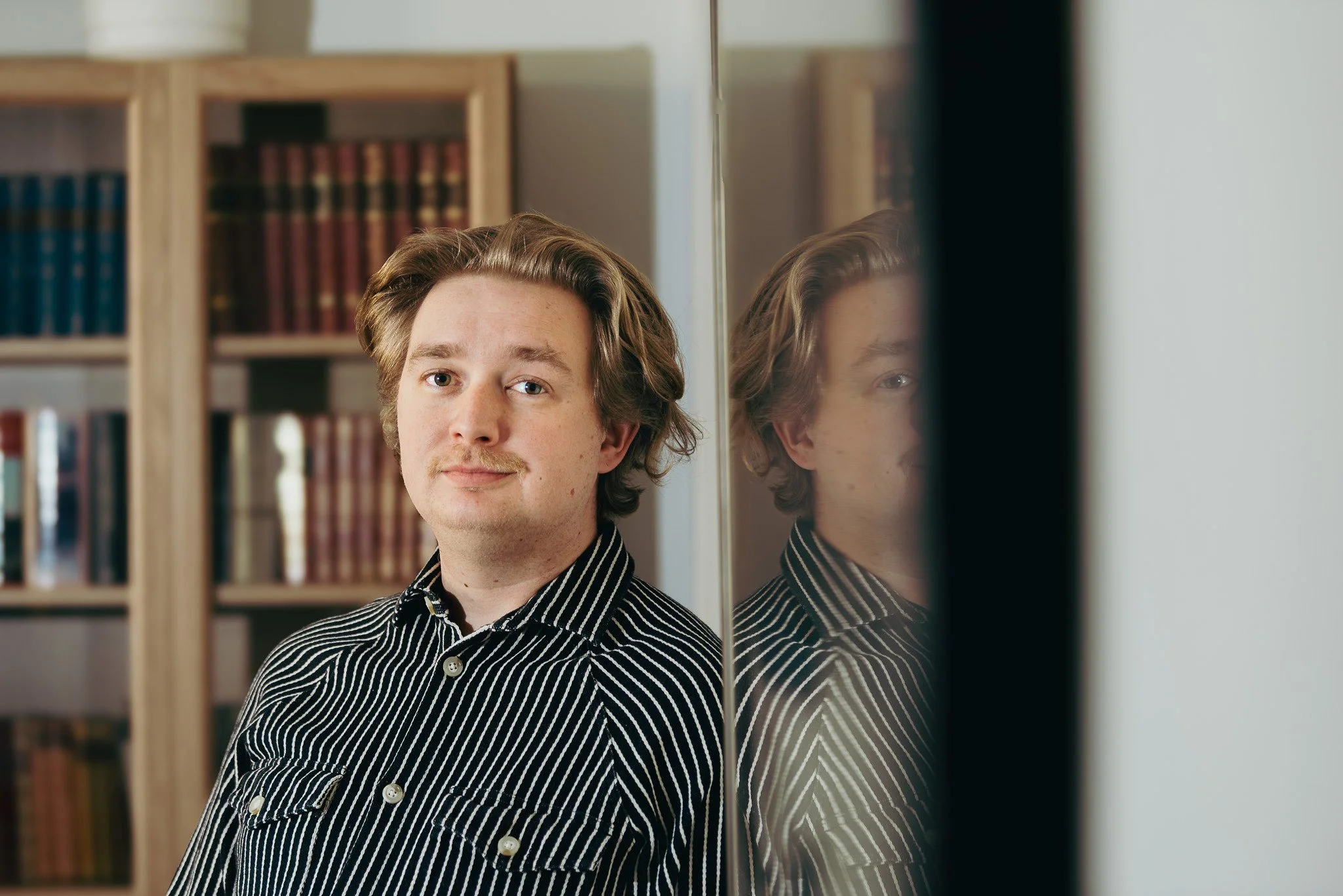 Raitapaitainen mies nojaa seinään ja katsoo kohti kameraa, takanaan kirjahyllyjä.