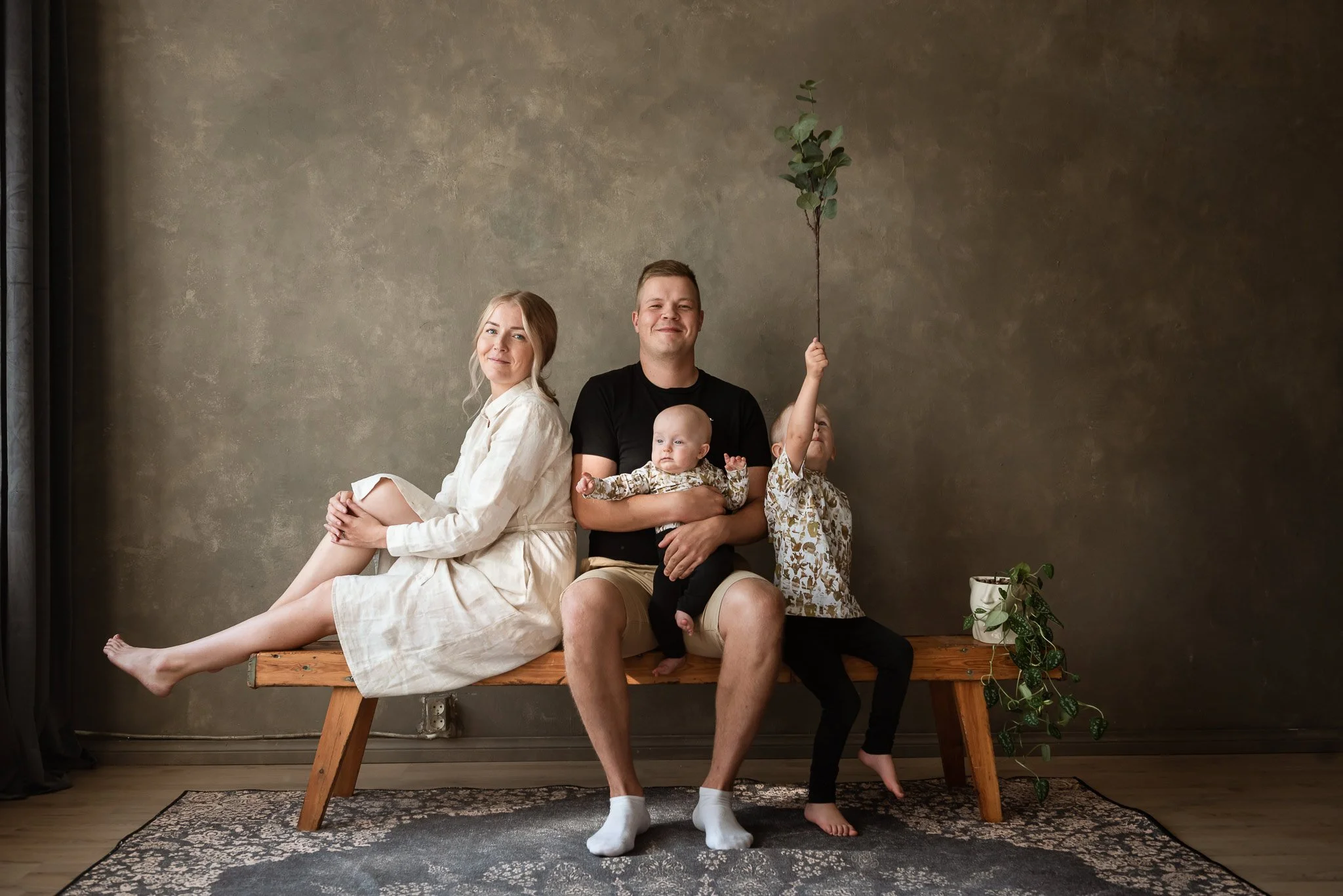 Maanläheisen ruskeansävyisessä studiossa puupenkillä istuu nelihenkinen perhe: äiti, isä, jolla on vauva sylissään sekä pieni poika, joka pitelee ylösojentuneessa kädessään eukalyptuksen oksaa.