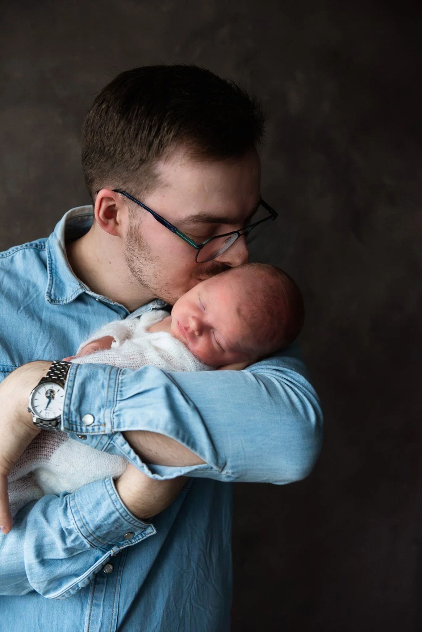 Siniseen farkkukankaiseen paitaan pukeutunut mies suukottaa sylissään nukkuvaa vauvaa.