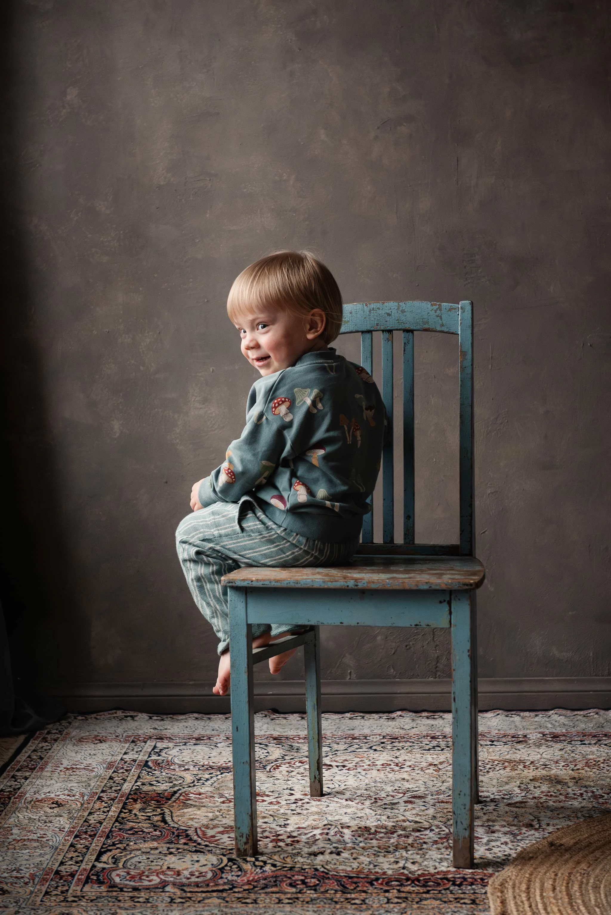 Olkansa yli katsova lapsi istuu sinisellä puutuolilla ja hymyilee.