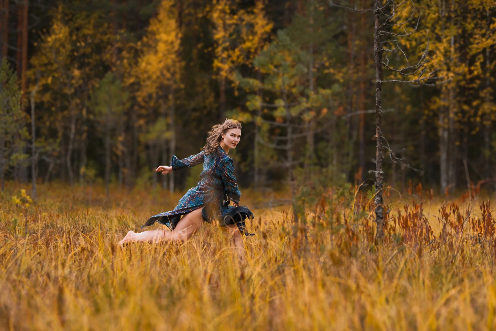 Tummansiniturkoosiin mekkoon pukeutunut nainen juoksee hiukset auki ja paljain jaloin syksyisessä suomaisemassa, takanaan tiheä metsä.