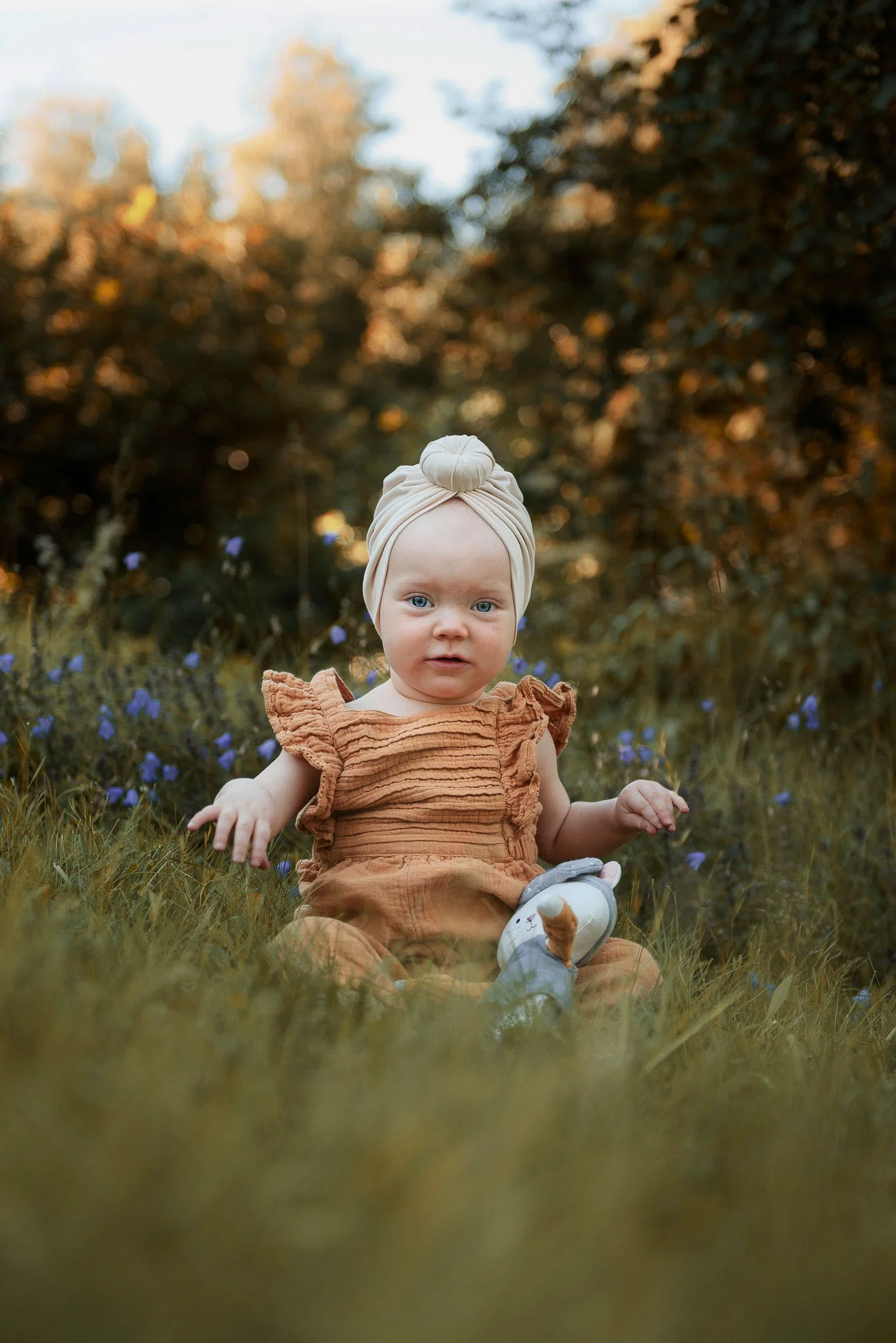 Murretun vaaleanoranssiin haalariin puettu taapero valkoinen huivi päässään istuu kukkaniityllä ilta-auringossa.