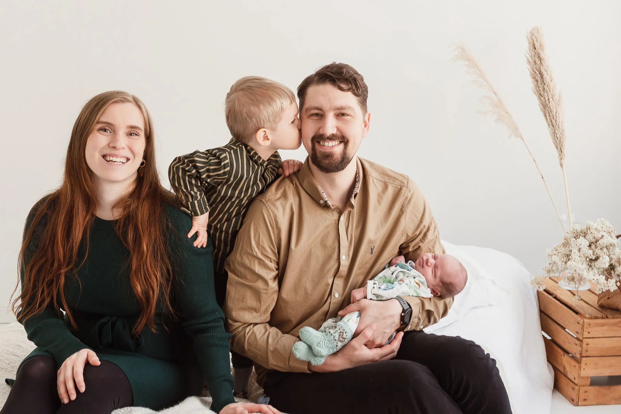 Valoisassa huoneessa valkoisella sängyllä istuu nelihenkinen perhe: hymyilevät äiti ja isä, jolla on vauva sylissään, sekä vanhempien välissä seisova pikkupoika, joka antaa isälleen pusun poskelle.