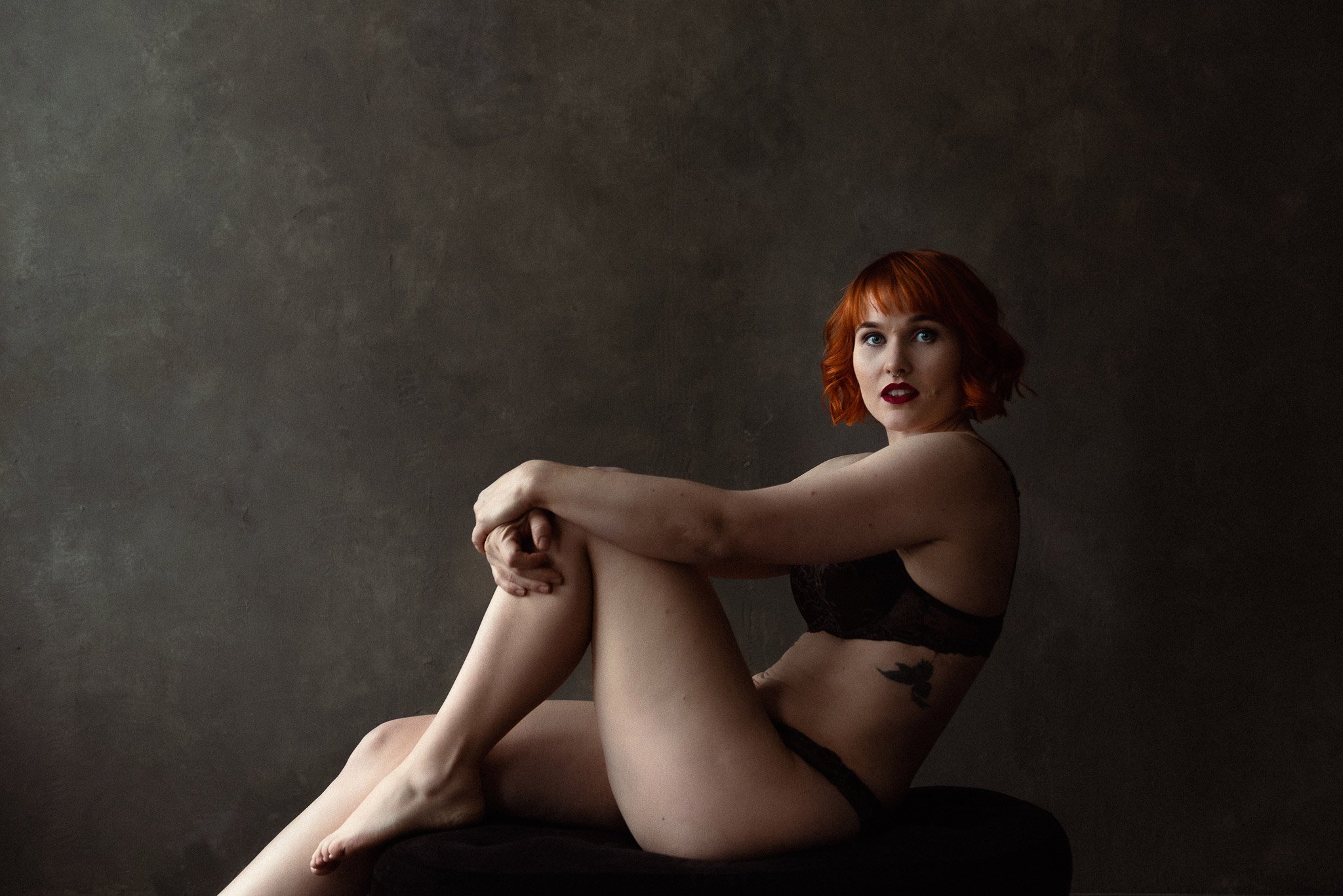 Punatukkainen, tummiin alusvaatteisiin pukeutunut nainen nojaa taaksepåin, käsivarret koukistetun polven ympärille kiedottuna.