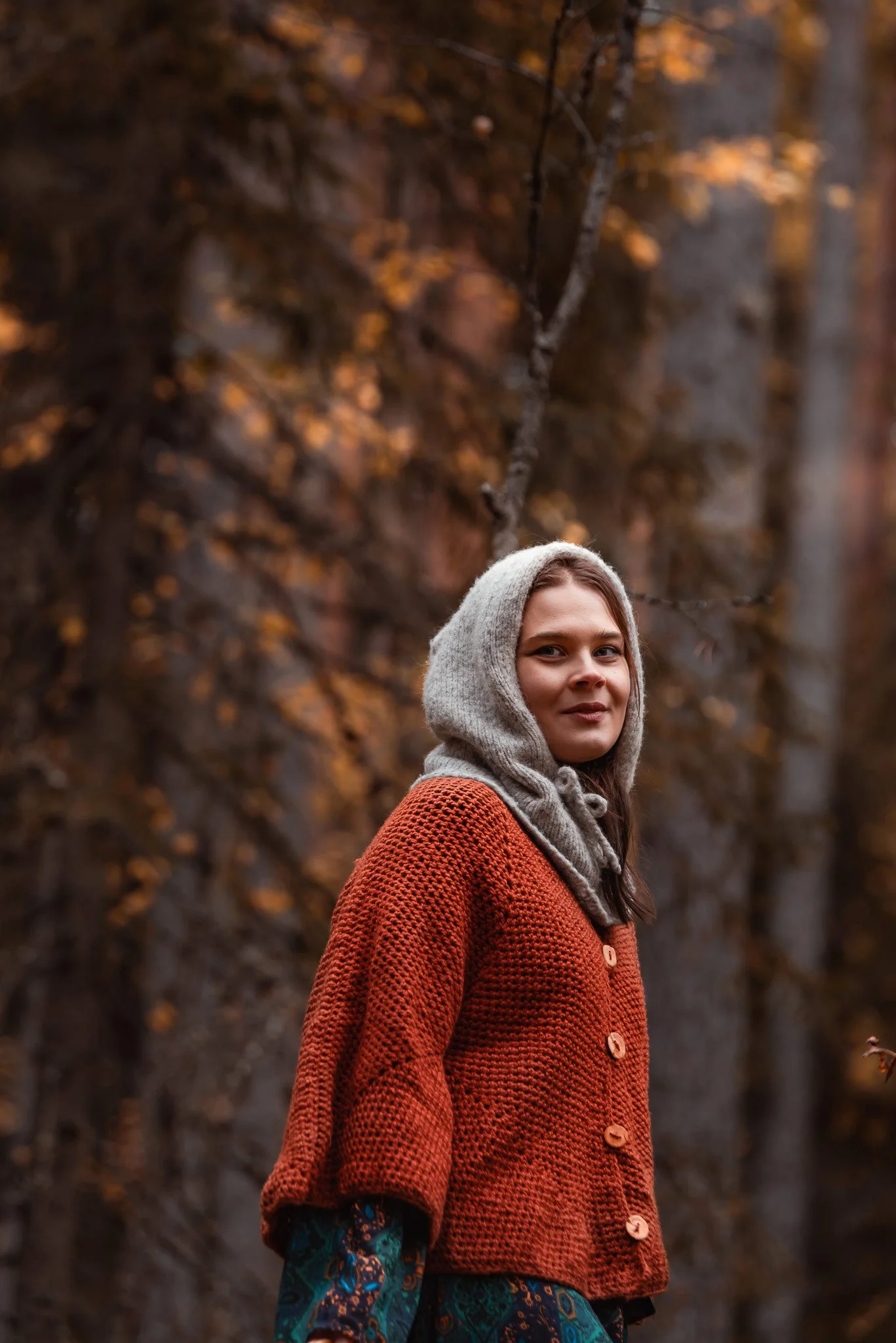 Punaoranssiin neuletakkiin ja harmaaseen villahuppuun pukeutunut nainen seisoo syksyisessä metsässä.