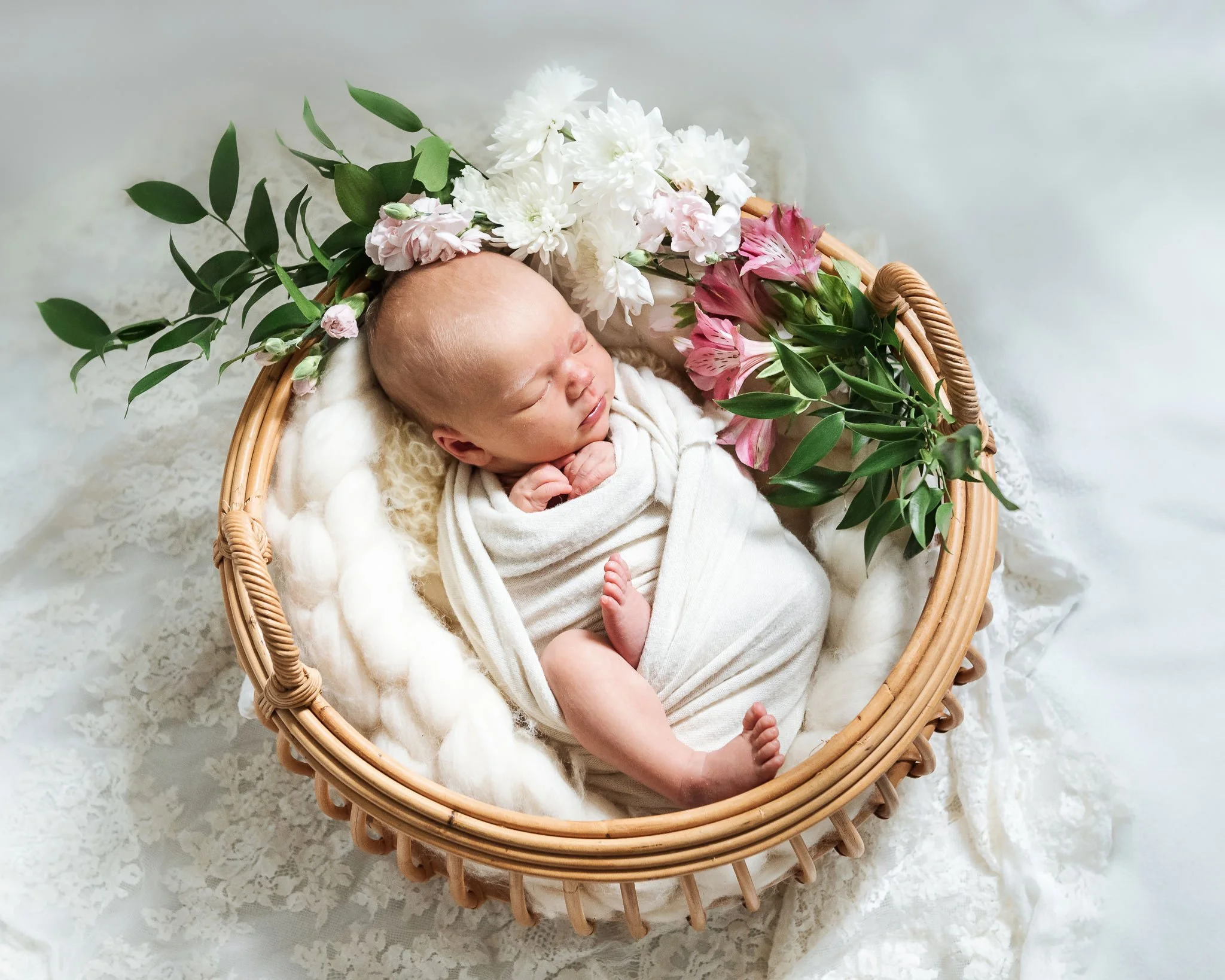 Valkoiseen vilttiin kääritty vauva nukkuu valkoisten ja vaaleanpunaisten kukkien ympäröimänä.