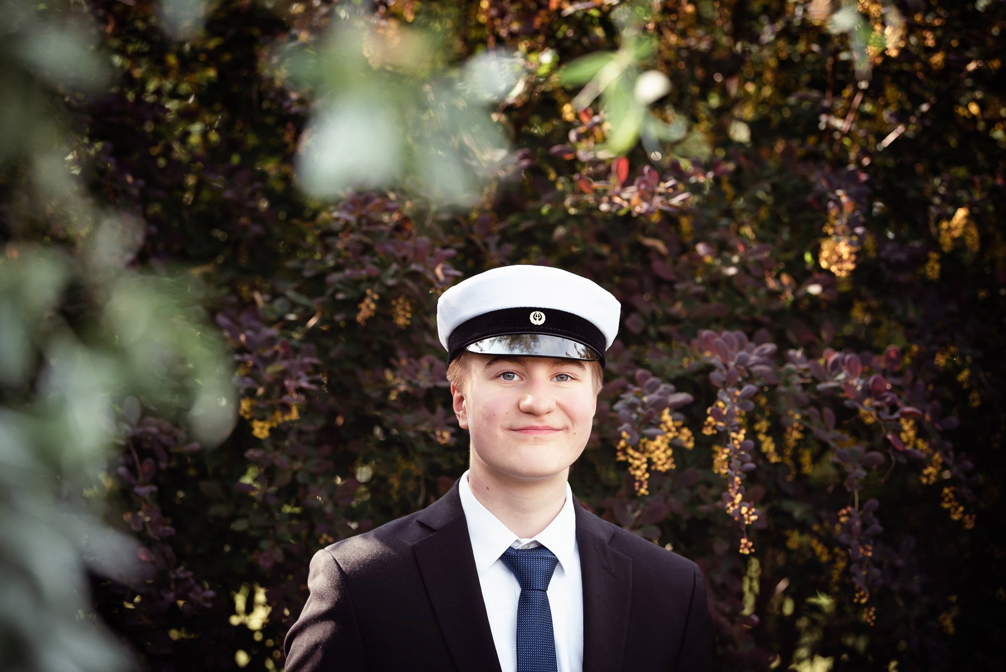 Nuorimies ylioppilaslakki päässään ja tummassa puvussa katsoo hymyillen kameraan, takanaan violetin ja keltaisen sävyisiä pensaita.