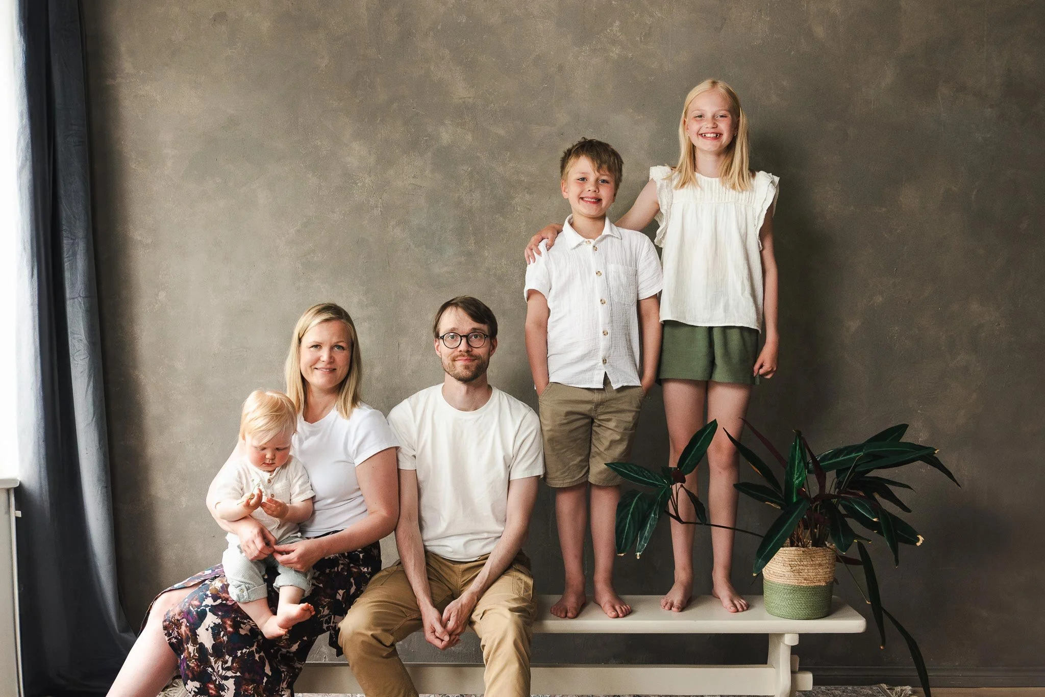 Maanläheisen ruskeansävyisen seinän edessä penkillä on viisihenkinen perhe: äiti, jolla on sylissään taapero, isä sekä penkillä seisovat alakouluikäiset lapset.