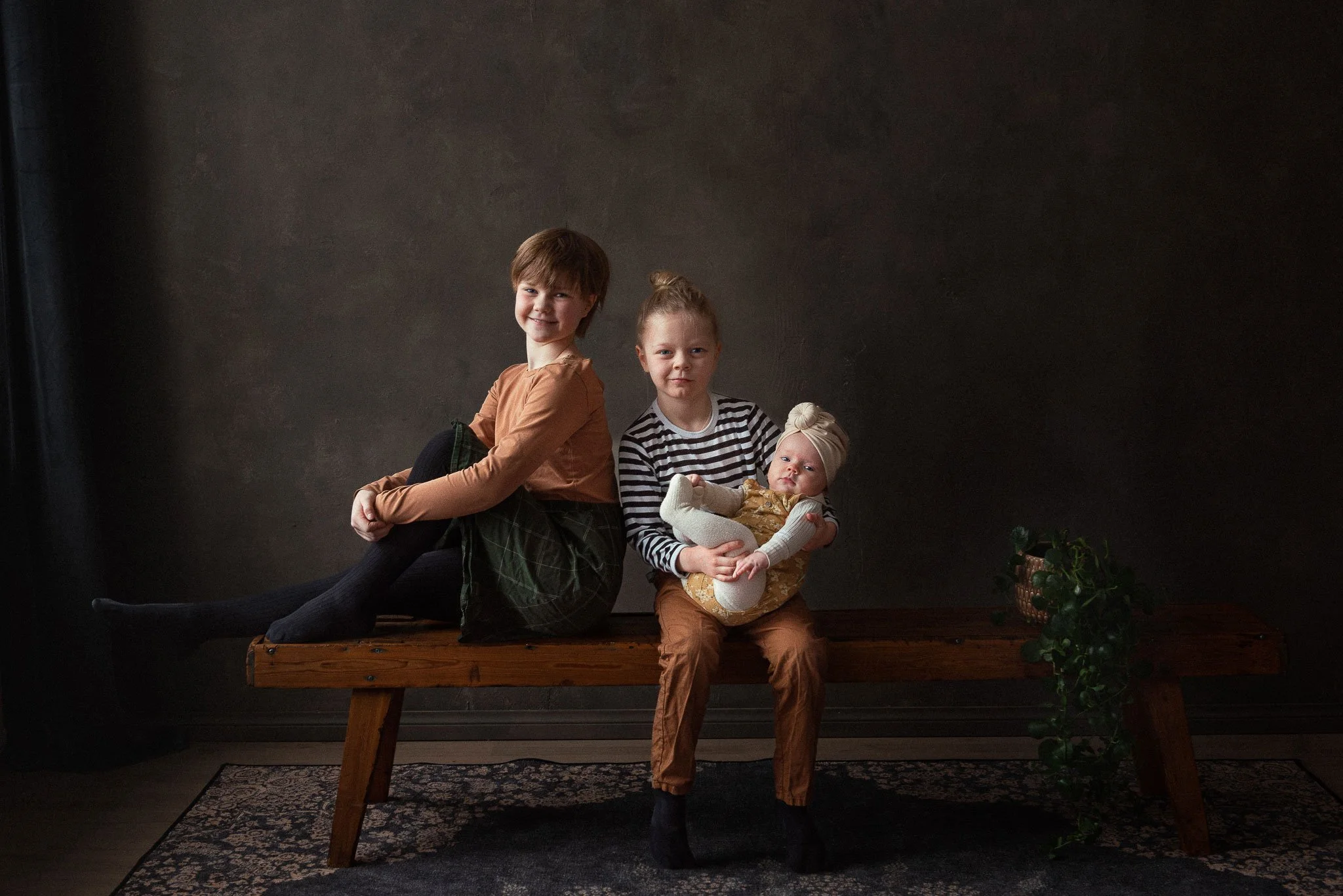 Tummasävyisessä studiossa istuu puupenkillä kaksi alakouluikäistä lasta, toinen pitelee sylissään vauvaa.
