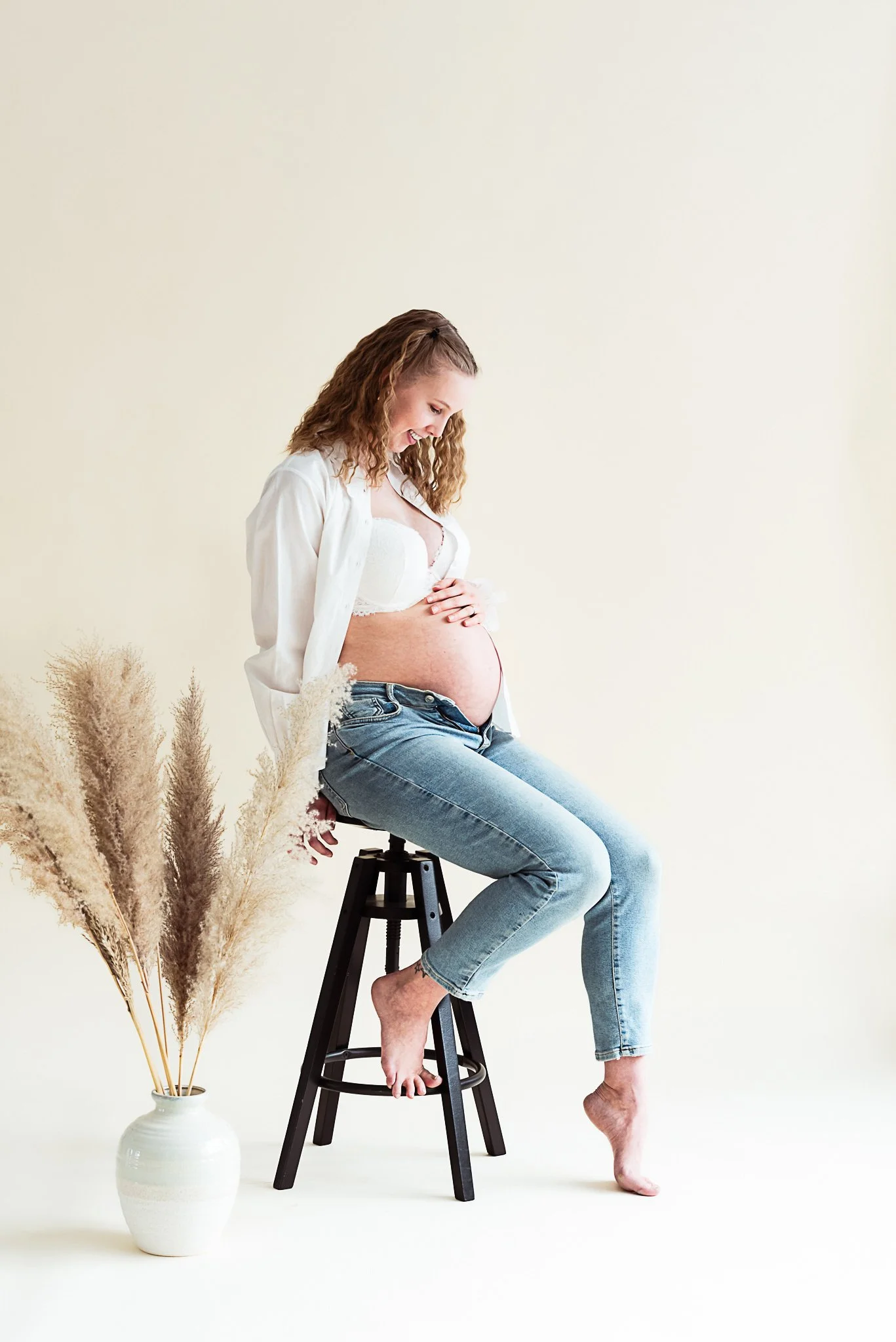 farkkuihin ja valkoiseen aukiolevaan kauluspaitaan pukeutunut raskaana oleva nainen istuu tuolilla valoisassa studiossa ja pitää kättään vatsansa päällä.