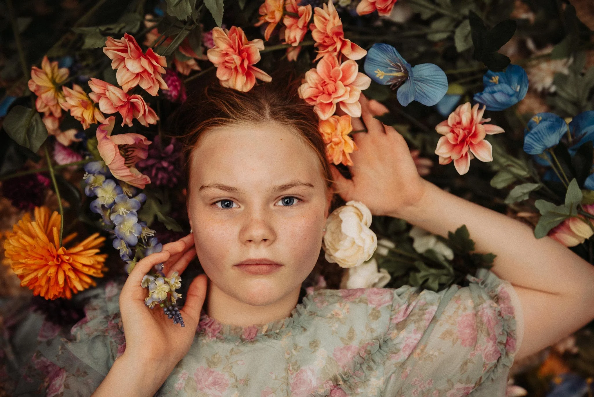 Alakouluikäinen lapsi makaa selällään oranssien, sinisten ja korallinpunaisten kukkien keskellä kädet lähellä kasvoja ja katsoo suoraan kameraan.