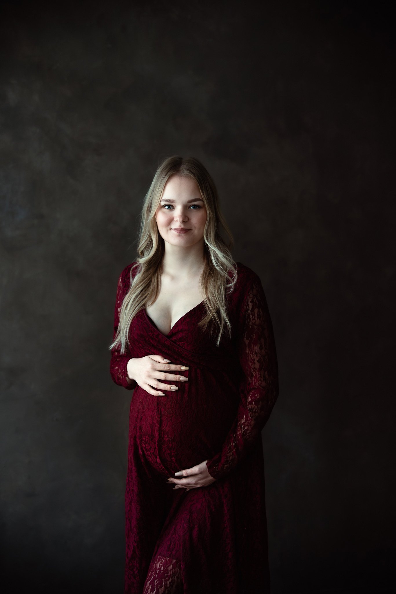 Burgundinpunaiseen pitsimekkoon pukeutunut nuori nainen katsoo hymyillen kameraan, käsien asento ja mekon laskokset tuovat esiin raskausvatsan.