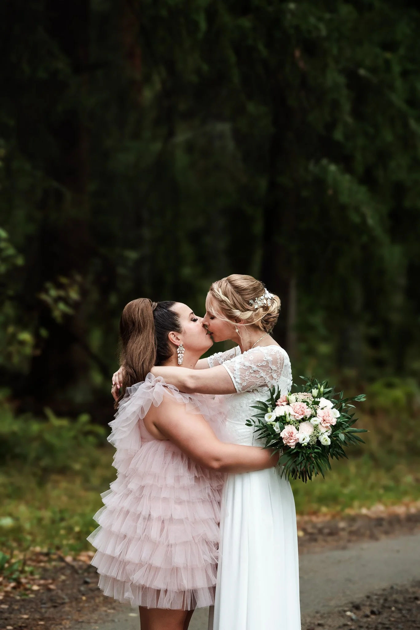 Valkoisiin ja vaaleanpunaisiin mekkoihin pukeutunut hääpari suutelee toisiaan tummassa kuusimetsässä.
