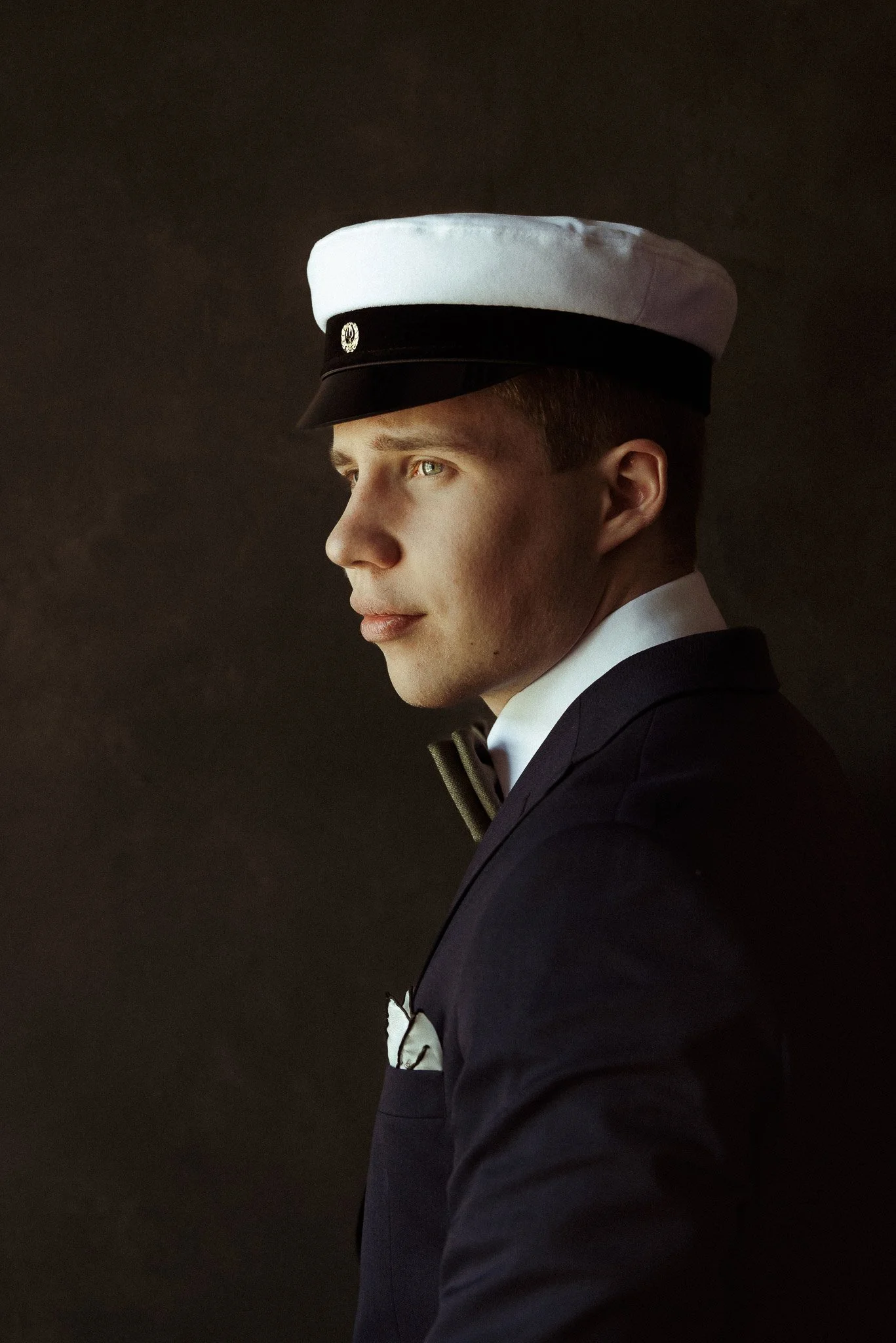 Sivuprofiilista kuvatun, ylioppilaslakkiin ja tummaan pukuun pukeutuneen nuoren miehen kasvoille lankeaa pehmeä valo muuten tummasävyisessä huoneessa.