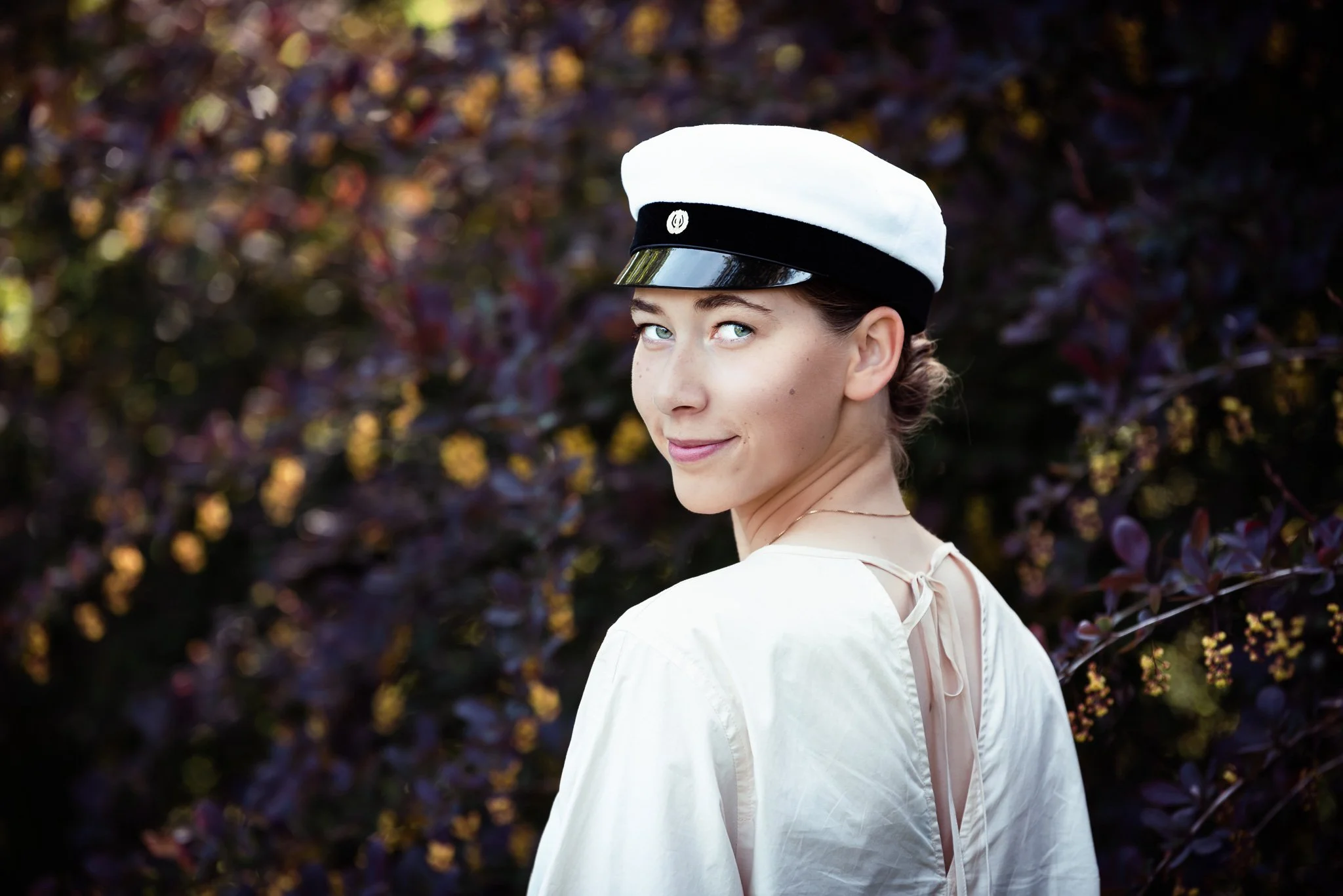 Valkoiseen mekkoon ja ylioppilaslakkiin pukeutunut nuori nainen katsoo olkansa yli kameraan, tummanviolettien pensaiden ympäröimänä.