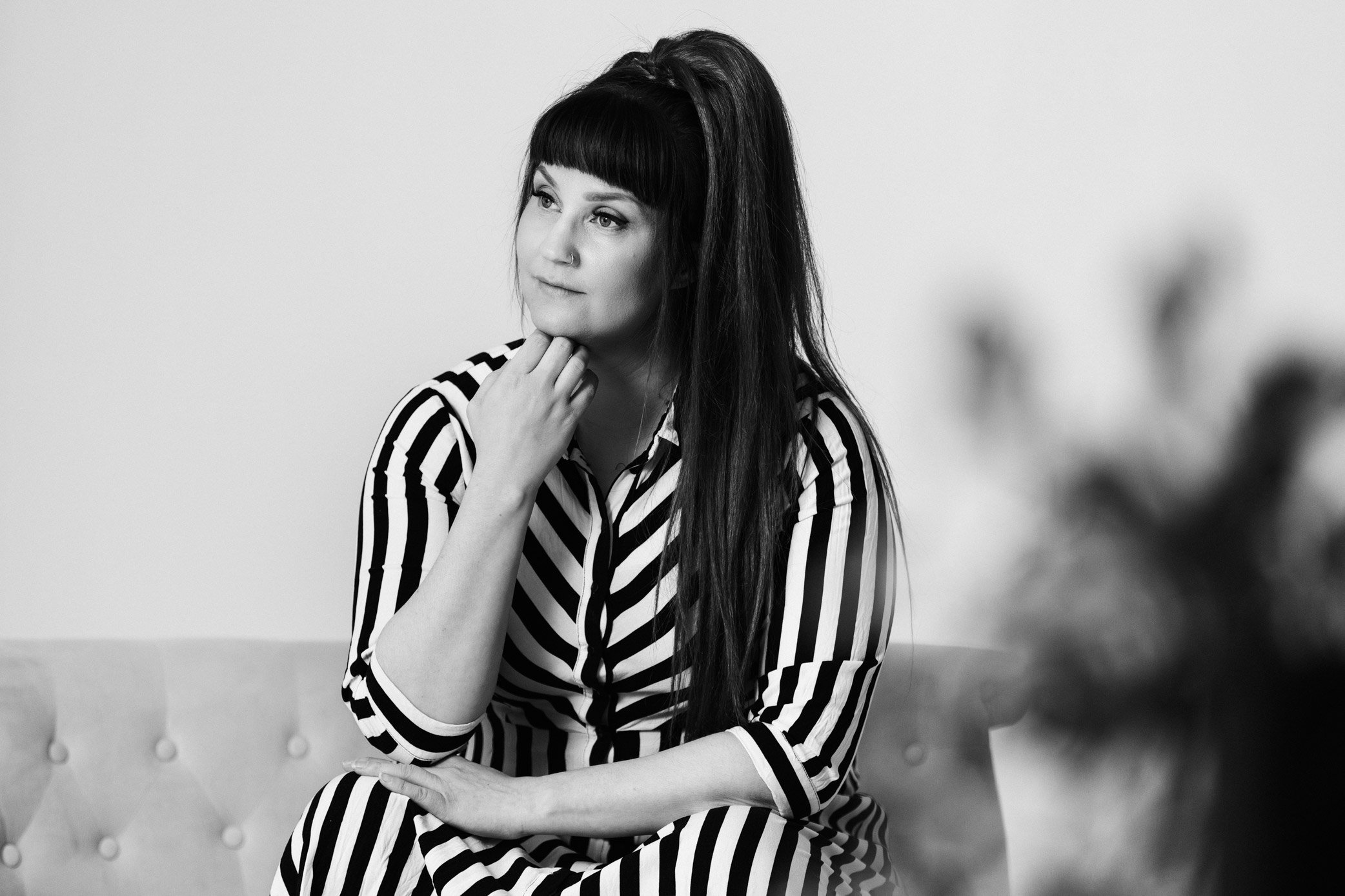 Raidalliseen mekkoon pukeutunut valokuvaaja Maarit Lahikainen istuu sohvan käsinojalla ja nojaa toista kättään leukaansa.