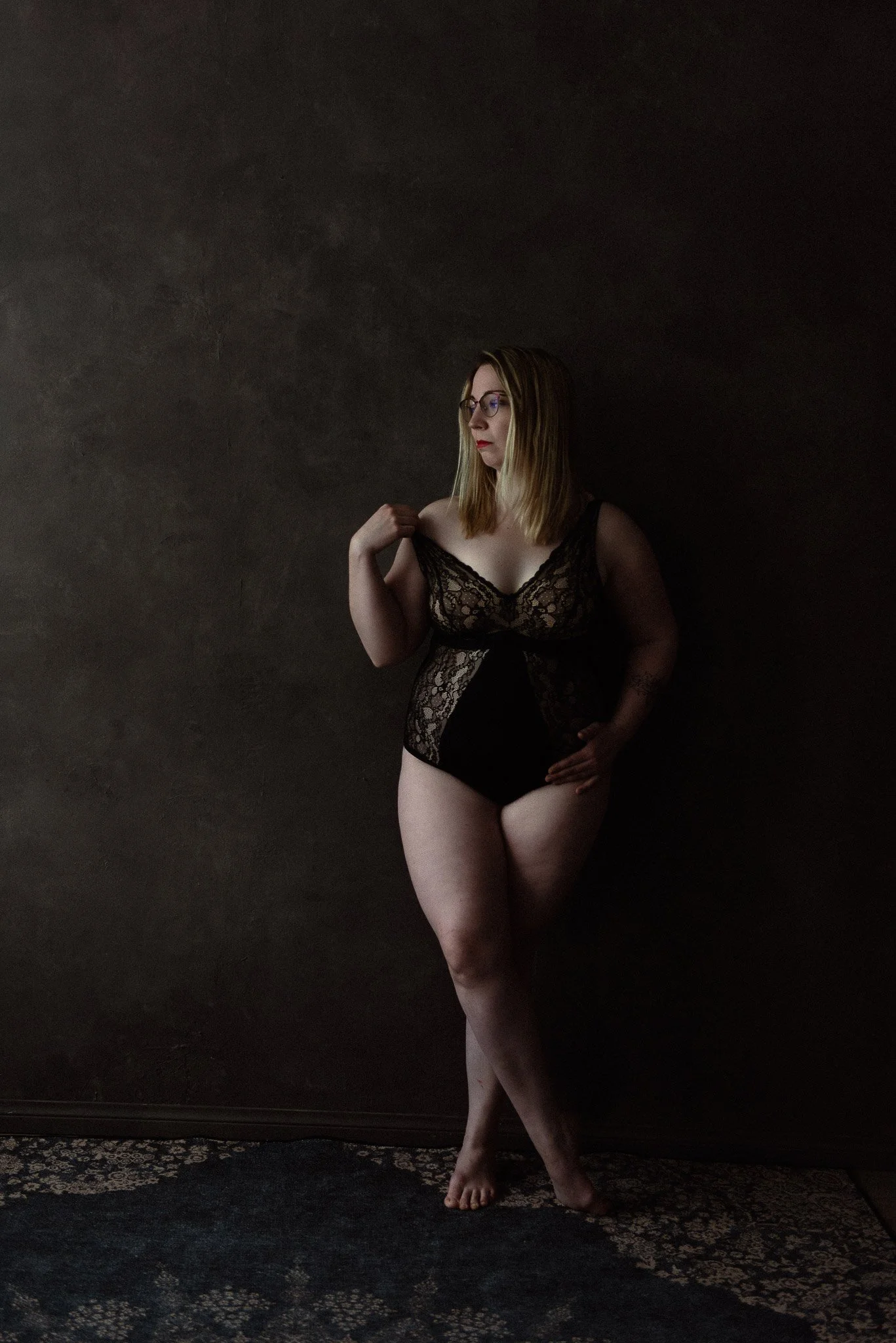 Mustaan pitsibodyyn pukeutunut silmälasipäinen nainen nojaa seinään, katse kääntyneenä vasemmalle..