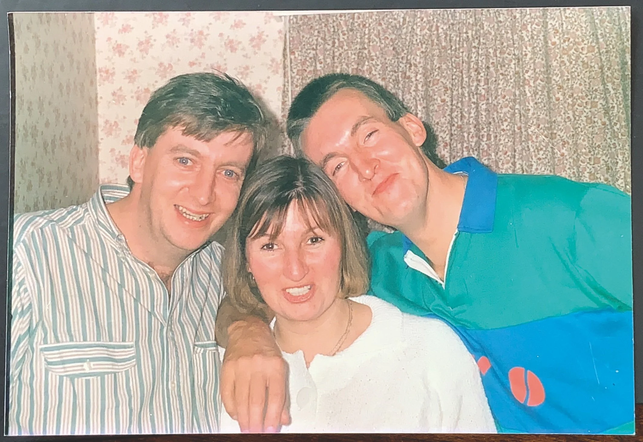 Three people smiling in a casual indoor setting, wearing casual attire. The background features floral wallpaper and a floral curtain.