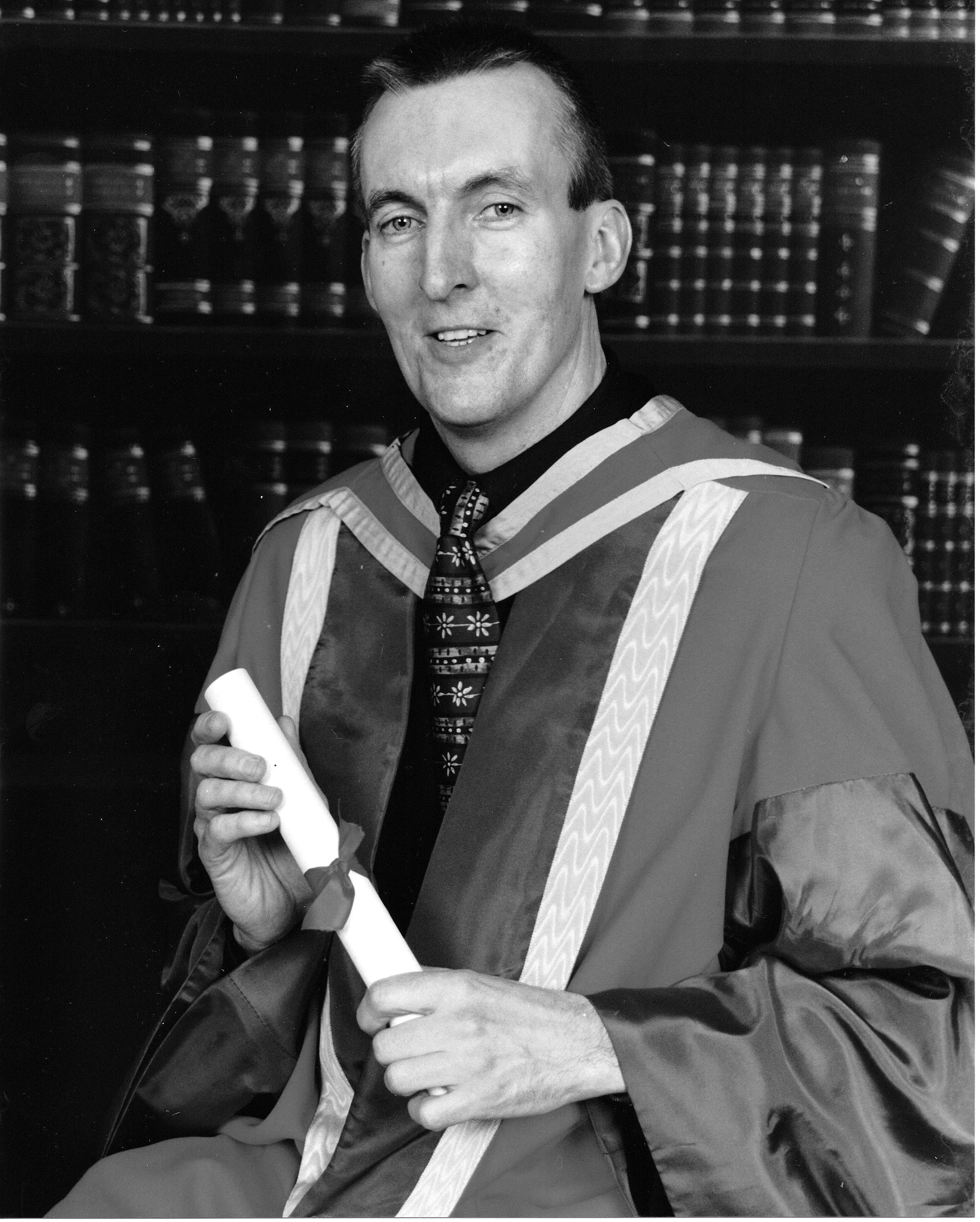 Person in graduation attire holding a diploma, black and white photo.