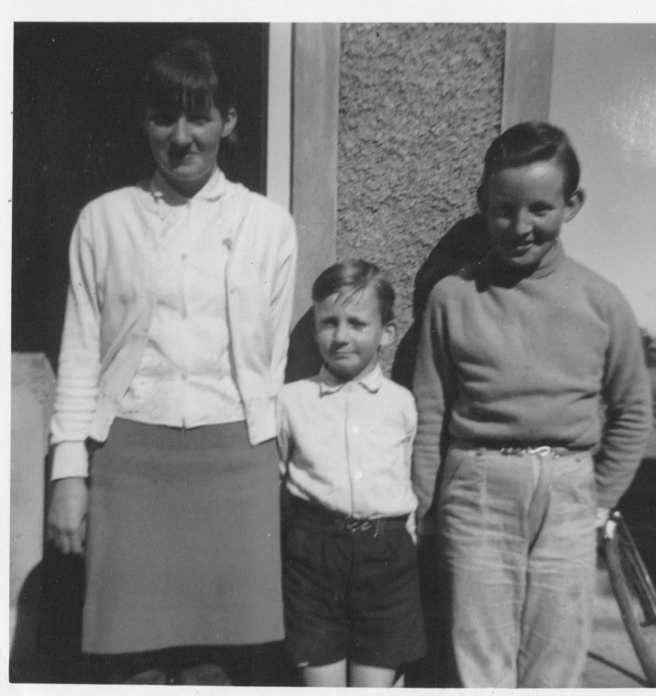 Three children, two boys and a girl, standing outside a building, smiling. The girl is wearing a cardigan and skirt, and the boys are in collared shirts and trousers. It's a black and white photo.
