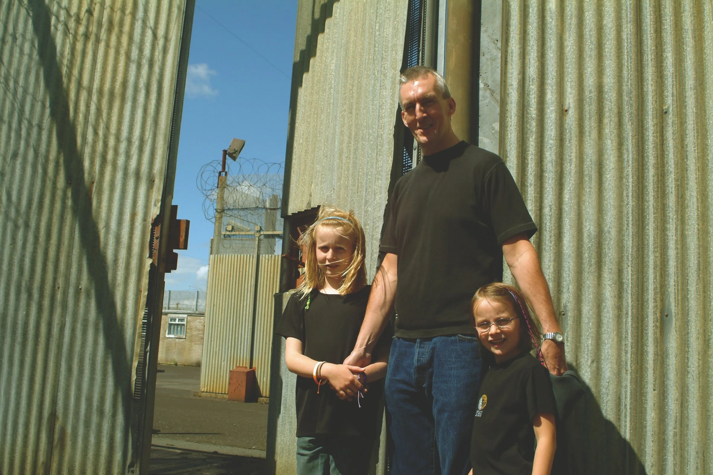 A man holding hands with two young girls, standing outdoors between corrugated metal walls.