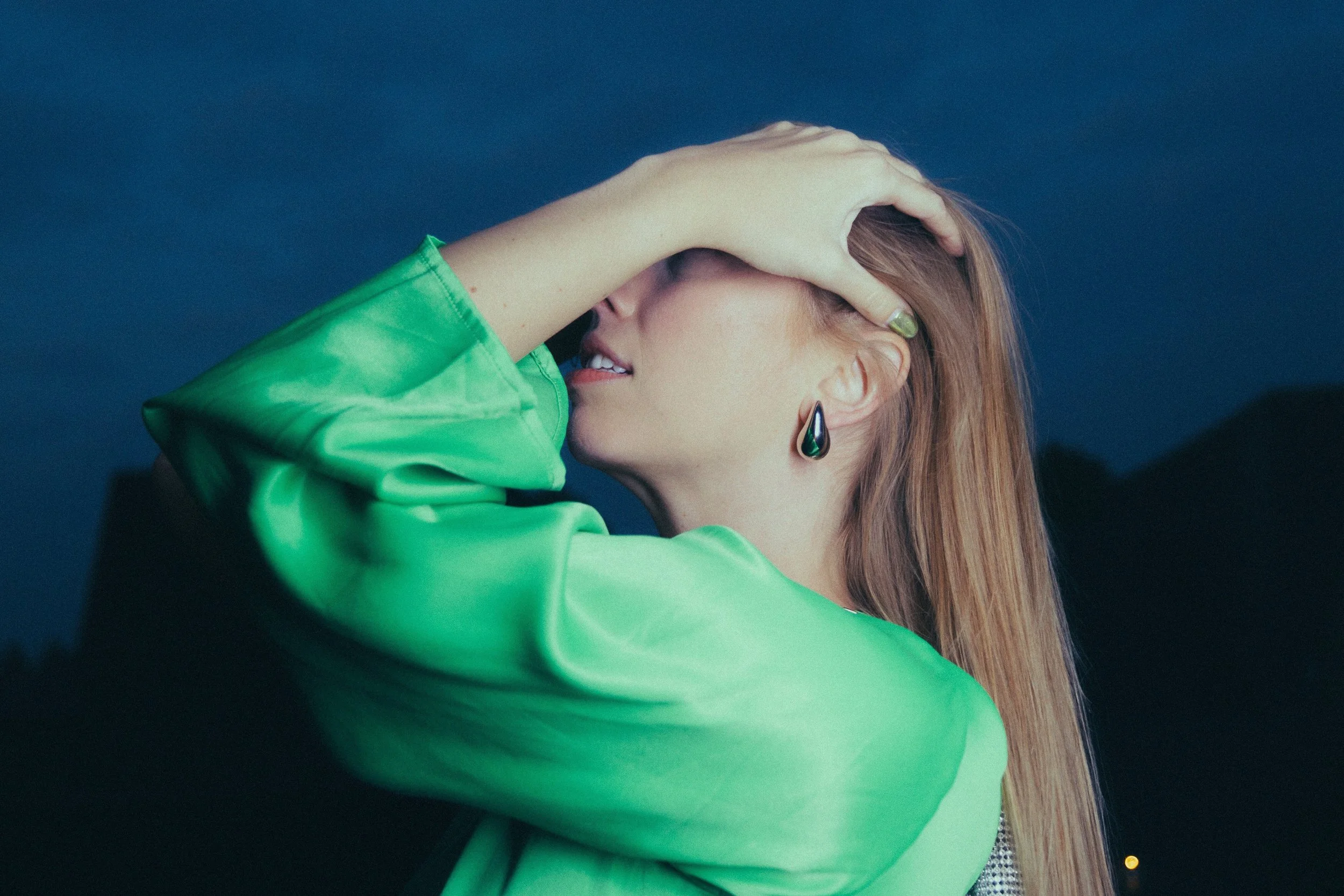 A woman with long reddish hair wearing a bright green jacket and black teardrop earrings, holding her head with one hand, against a dark blue evening sky.