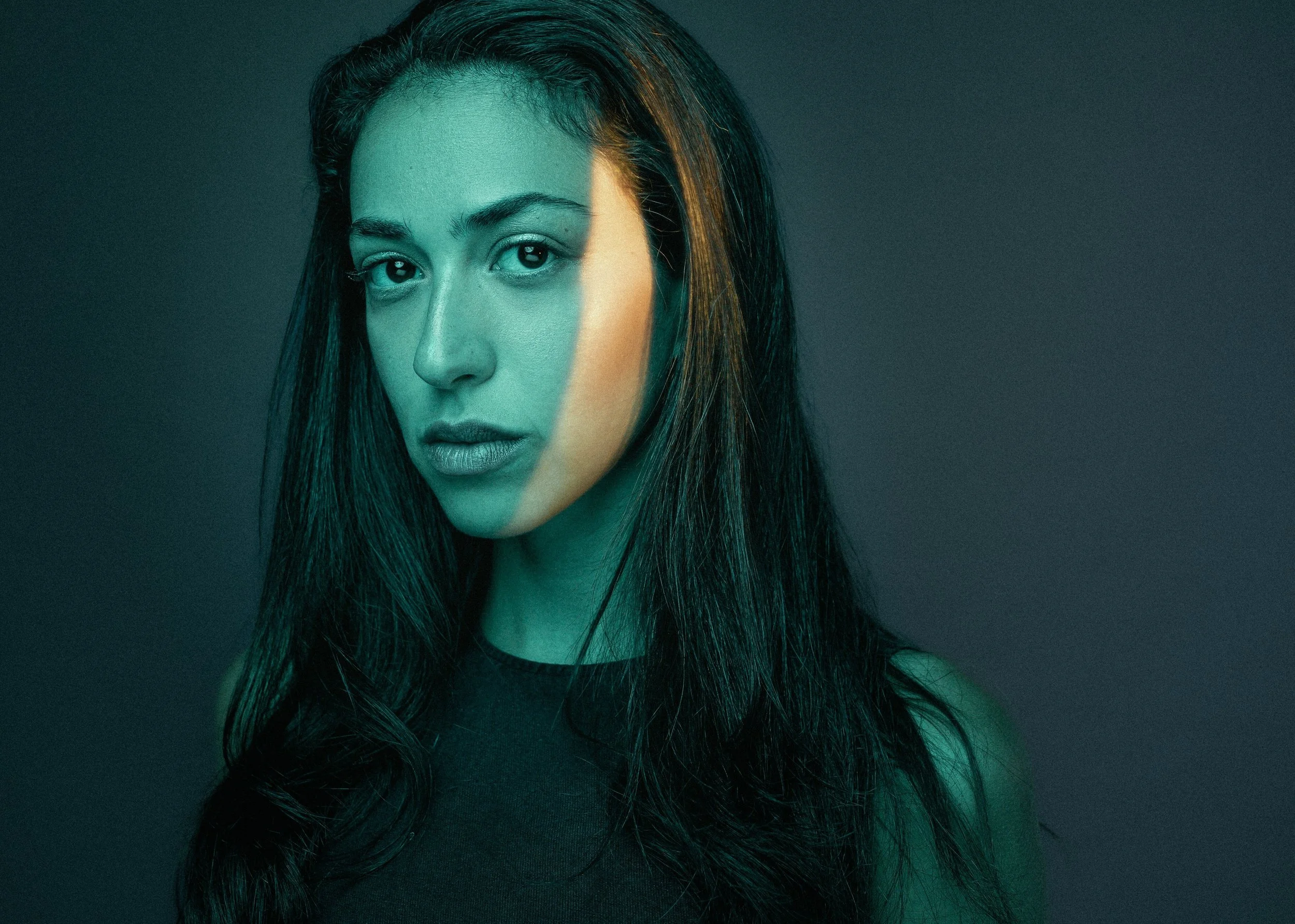 A woman with long dark hair stares at the camera with a serious expression against a dark background, her face partially illuminated with teal lighting.
