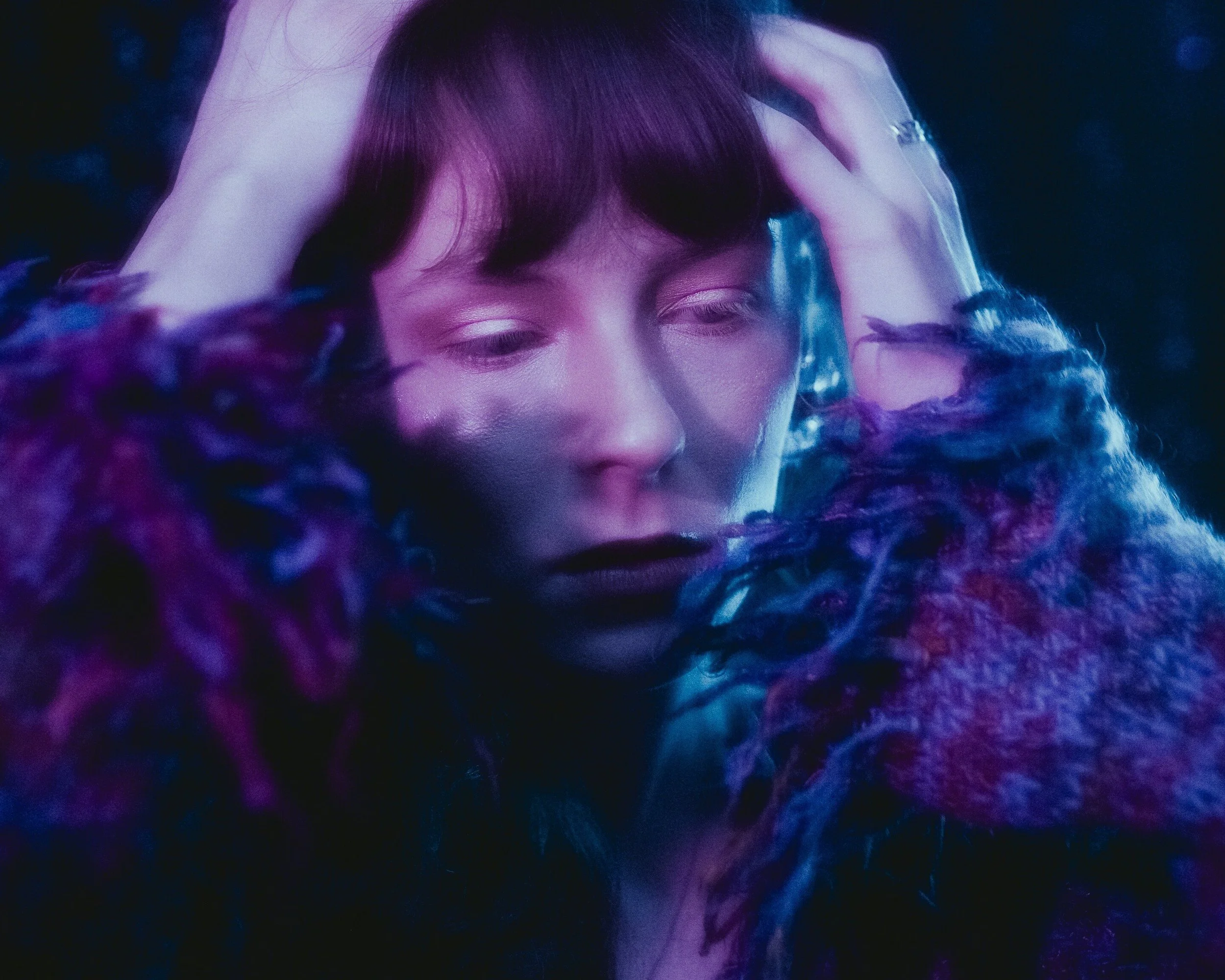 A person with short dark hair and light-colored eyes holds their head with both hands, with a contemplative expression, surrounded by colorful, textured fabrics in purple and blue hues.