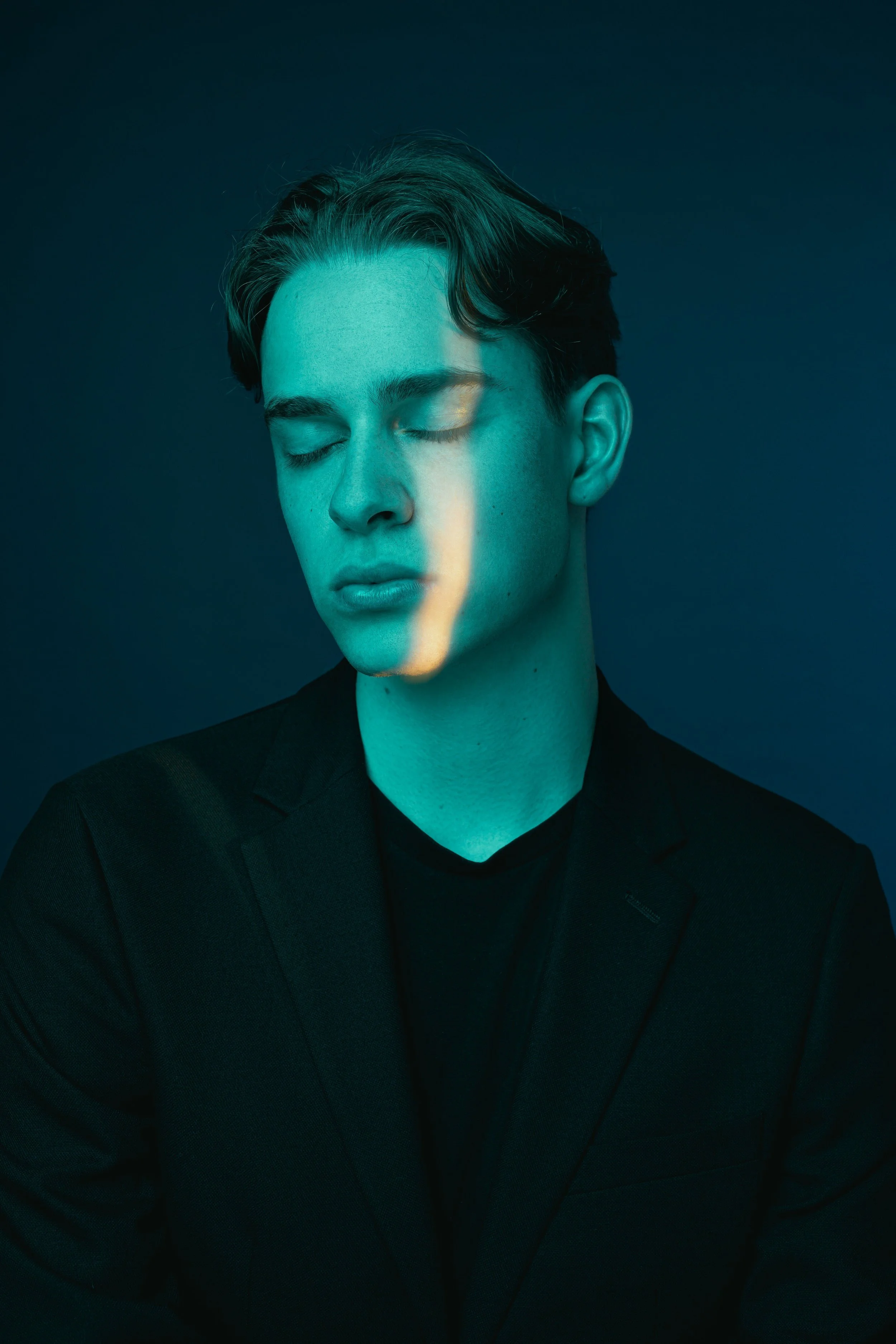 Portrait of a young man with closed eyes, light teal illumination on face, dark background, wearing a black blazer and shirt.