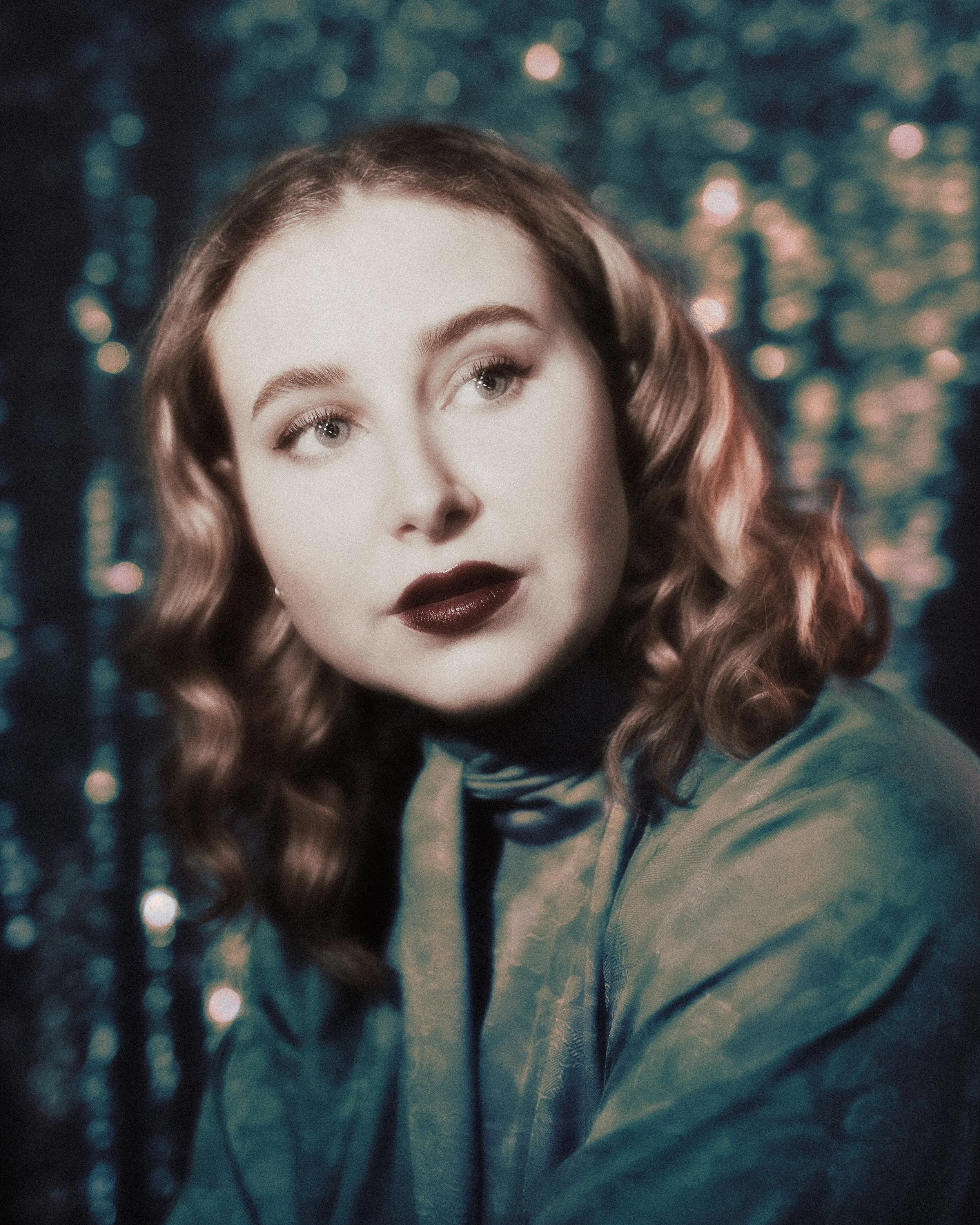 Portrait of a young woman with wavy brown hair and dark lipstick, wearing a blue dress, against a bokeh background of colorful lights.