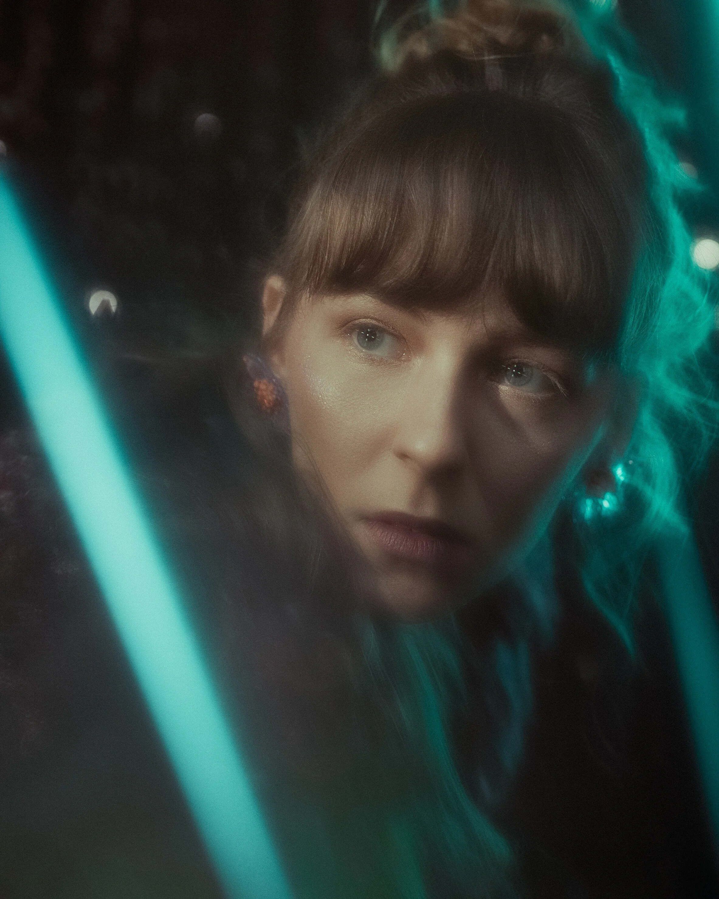 A young woman with blue eyes and brown hair looking through a glass with neon blue lights reflecting on her face.