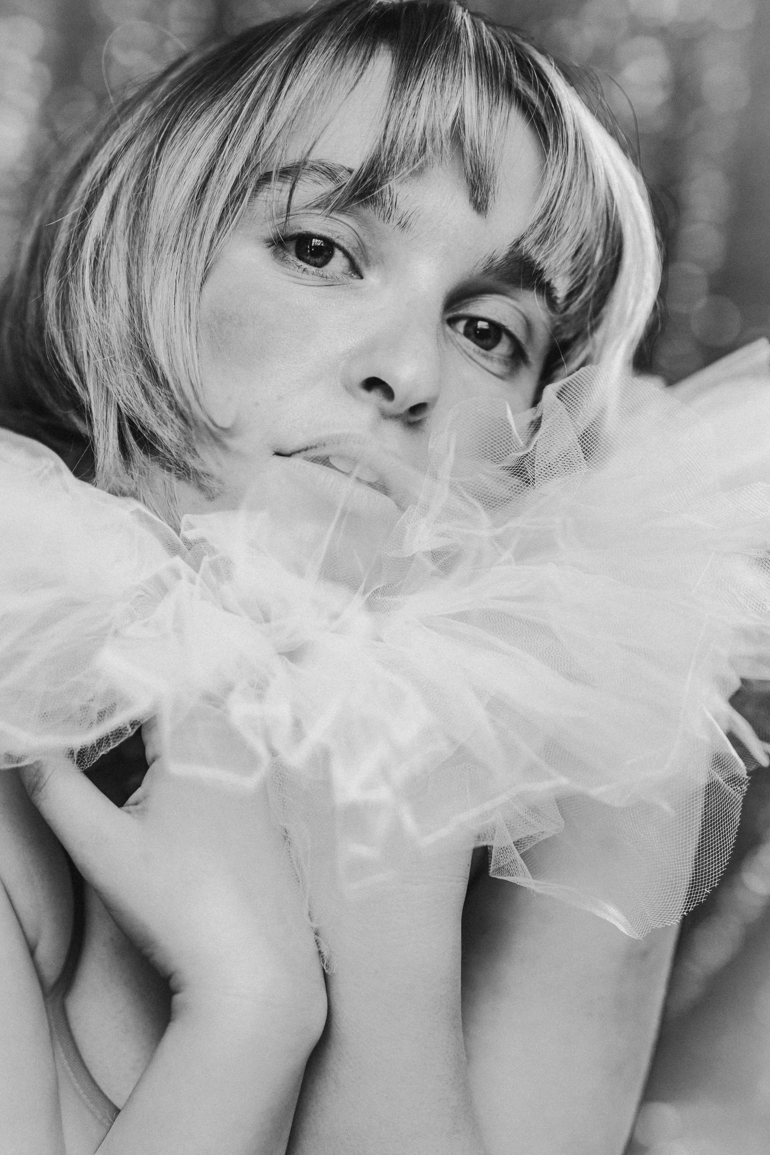 A black and white close-up portrait of a woman with short hair, gazing at the camera, with her hand near her face, wearing a large ruffled tulle accessory around her neck.