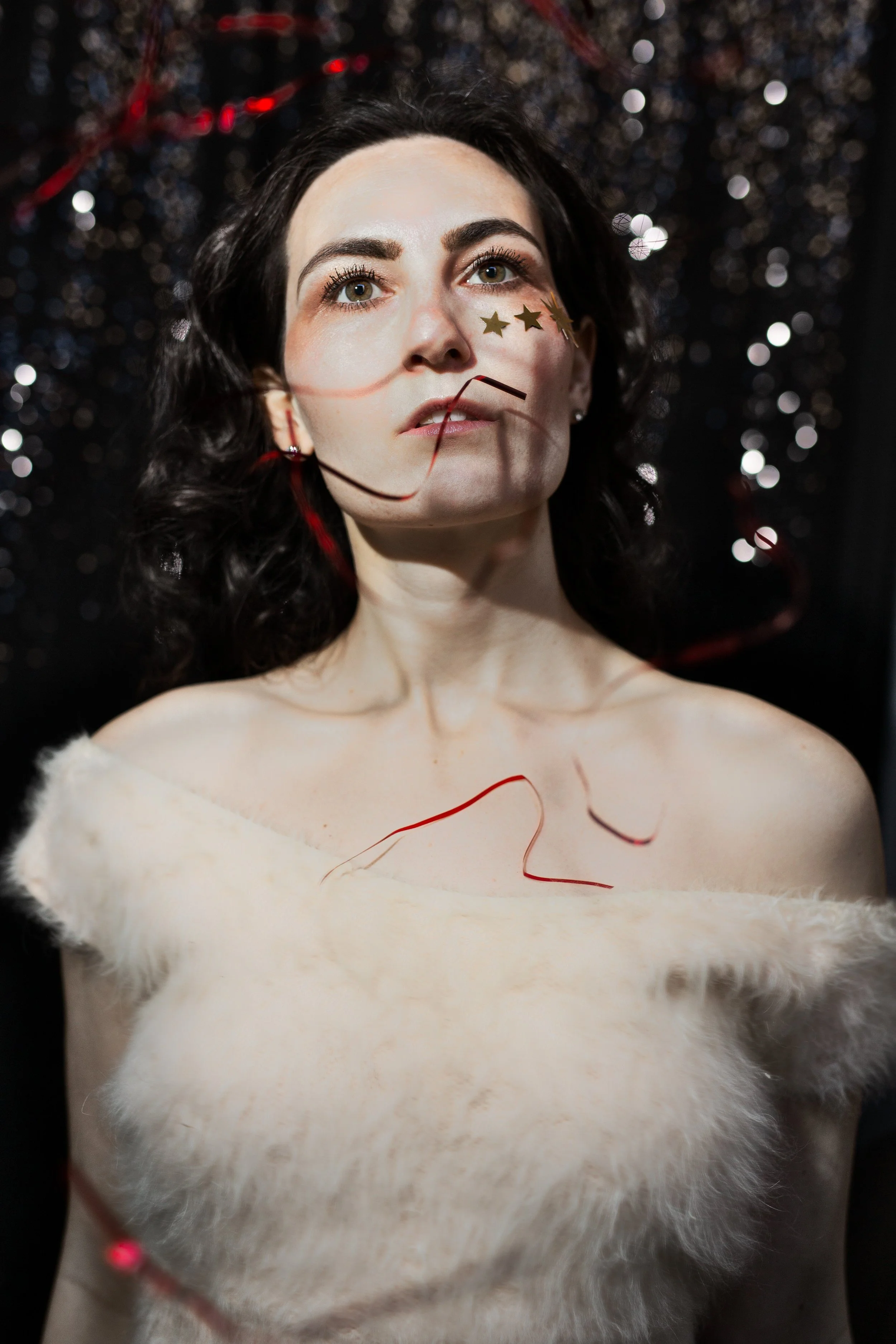 A woman with dark, curly hair and fair skin is wearing a white, fluffy off-shoulder top. She has star-shaped gold stickers under her right eye and some red string or ribbon across her face and body. The background is black with blurry silver and red 