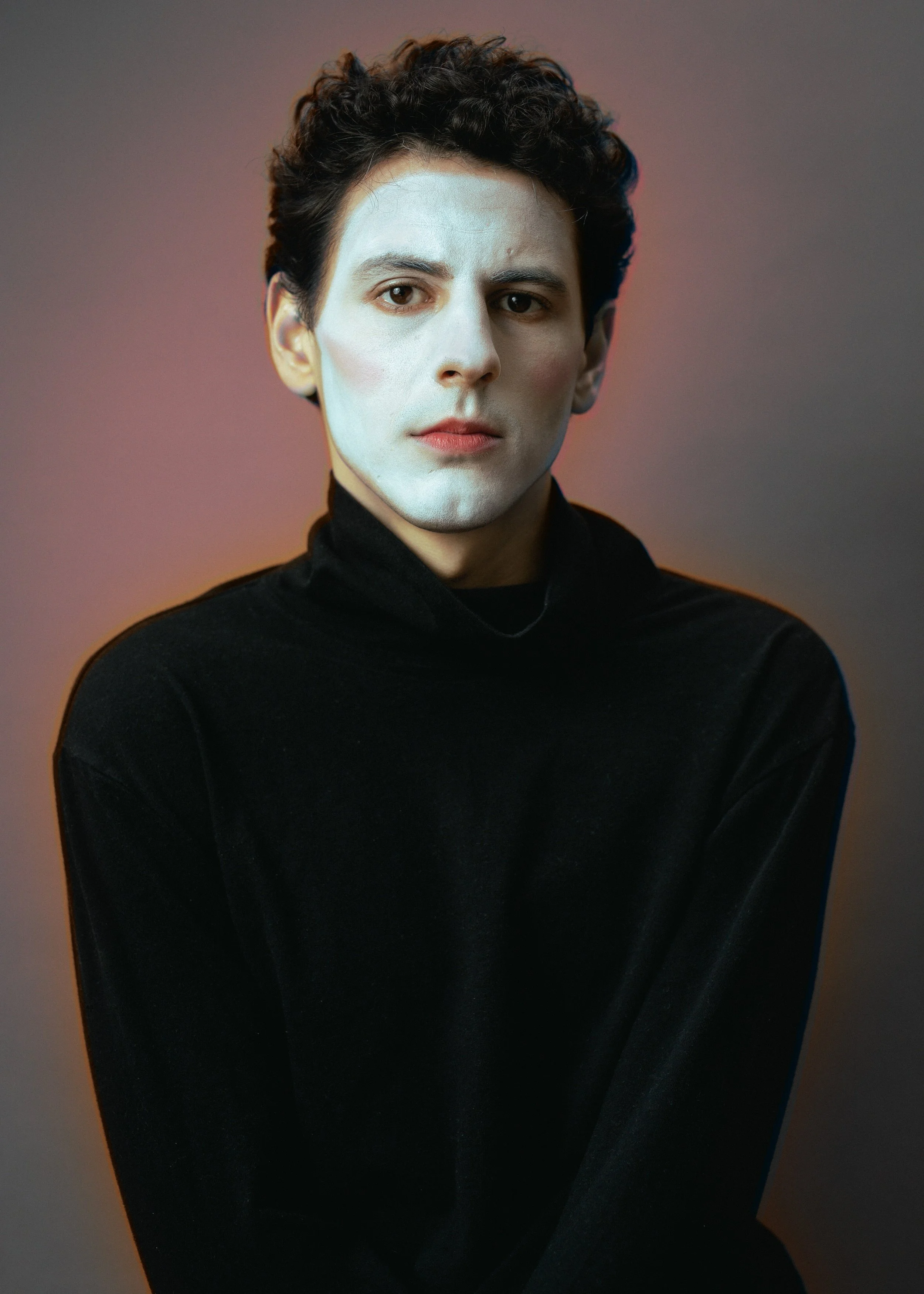 Portrait of a young man with curly dark hair and light makeup, wearing a black turtleneck, against a blurred gradient background.