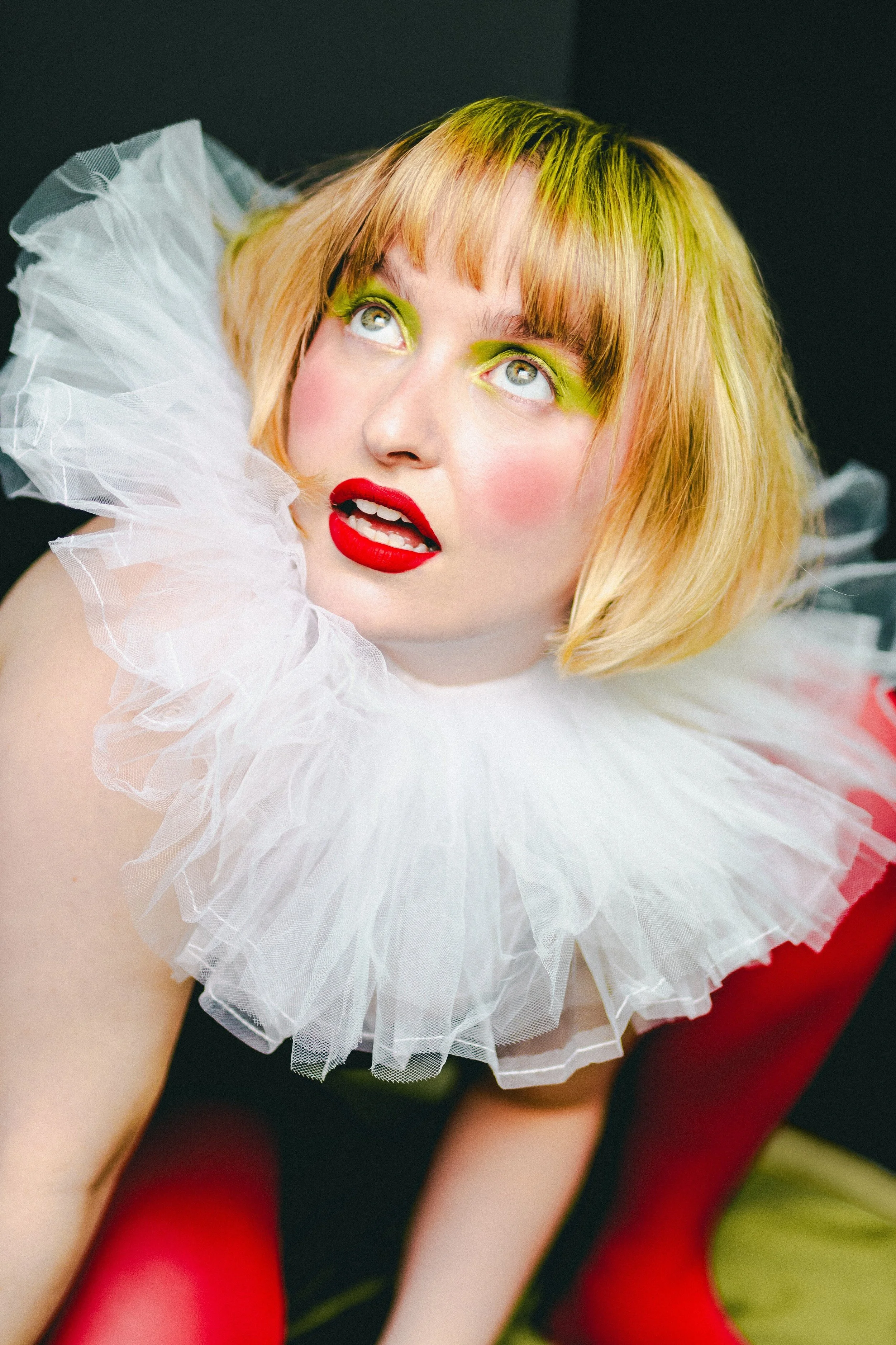A woman with blonde hair styled in a short bob, wearing bright green eyeshadow, bold red lipstick, and a white ruffled collar, looking upwards with a surprised expression.