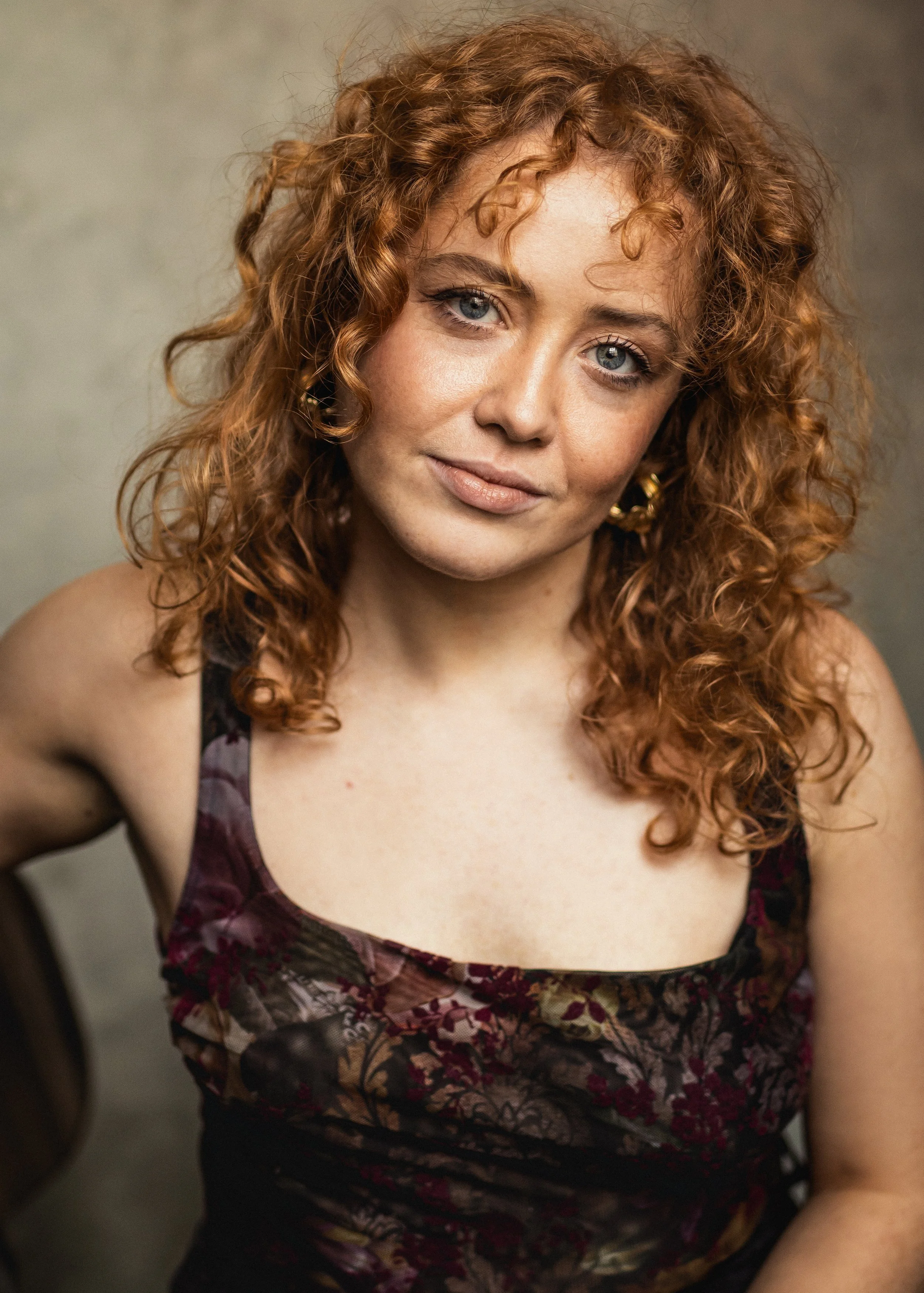 A young woman with curly red hair and blue eyes looking at the camera. She is wearing a floral dress with thin straps and gold hoop earrings.