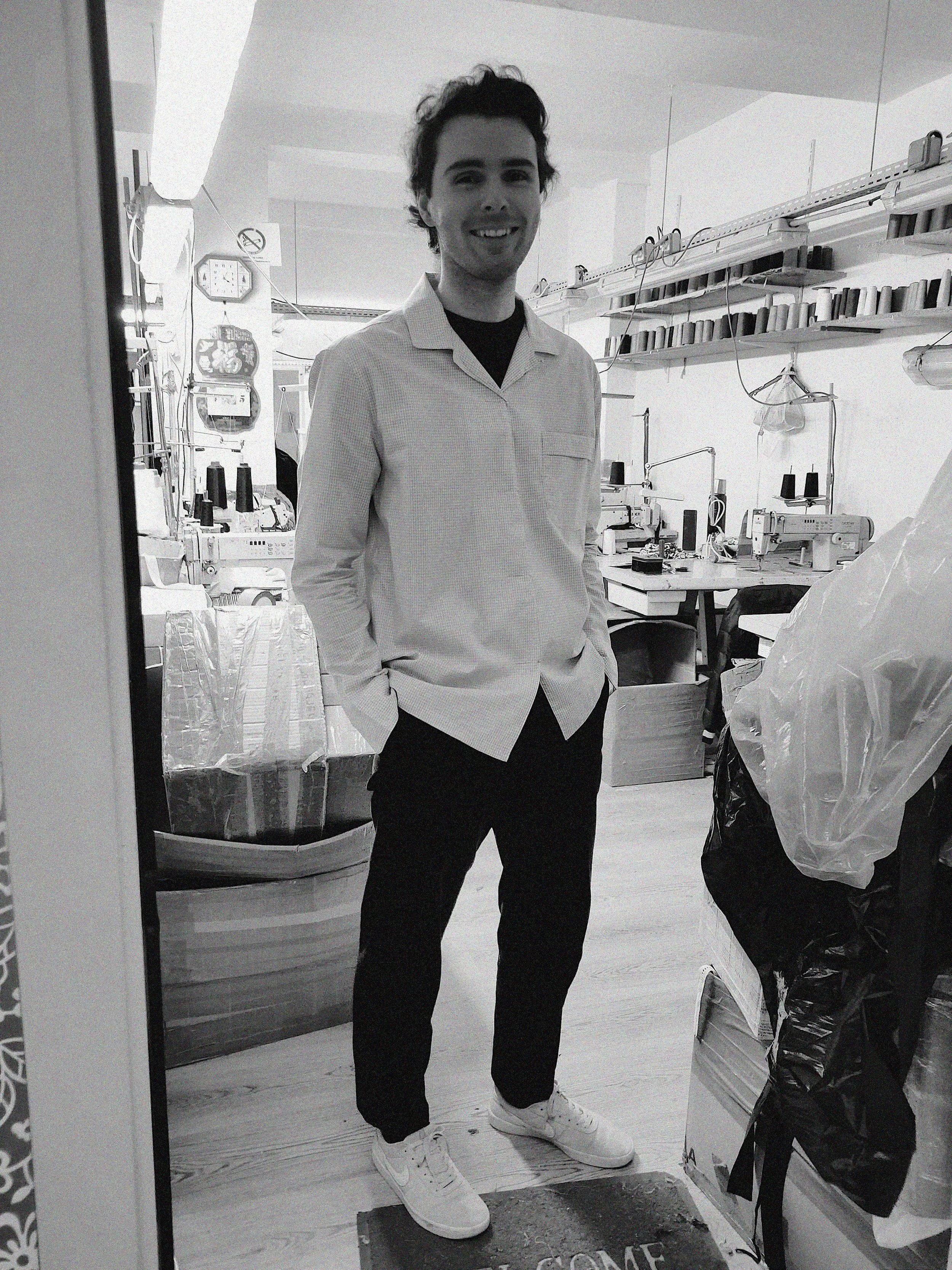 Un jeune homme souriant, en chemise claire, debout dans un atelier avec des machines à coudre, des bobines de fil, et des sacs en plastique autour.