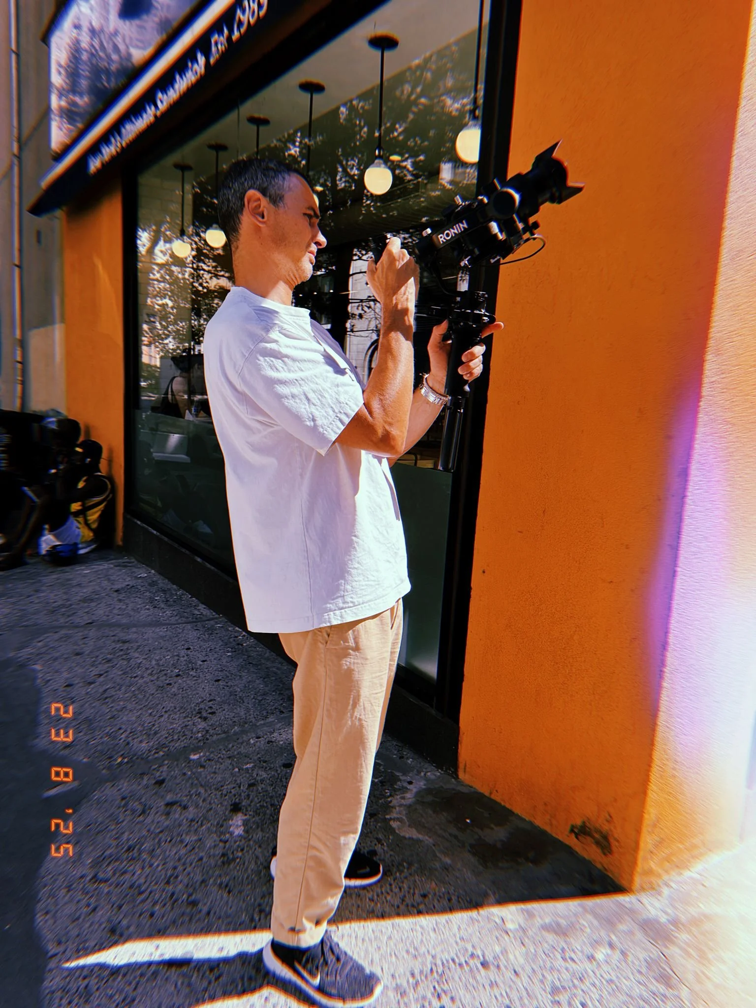 A man wearing a white T-shirt and beige pants is standing outside, holding a camera on a stabilizer, aiming it towards a storefront window with reflections of trees and hanging lights.