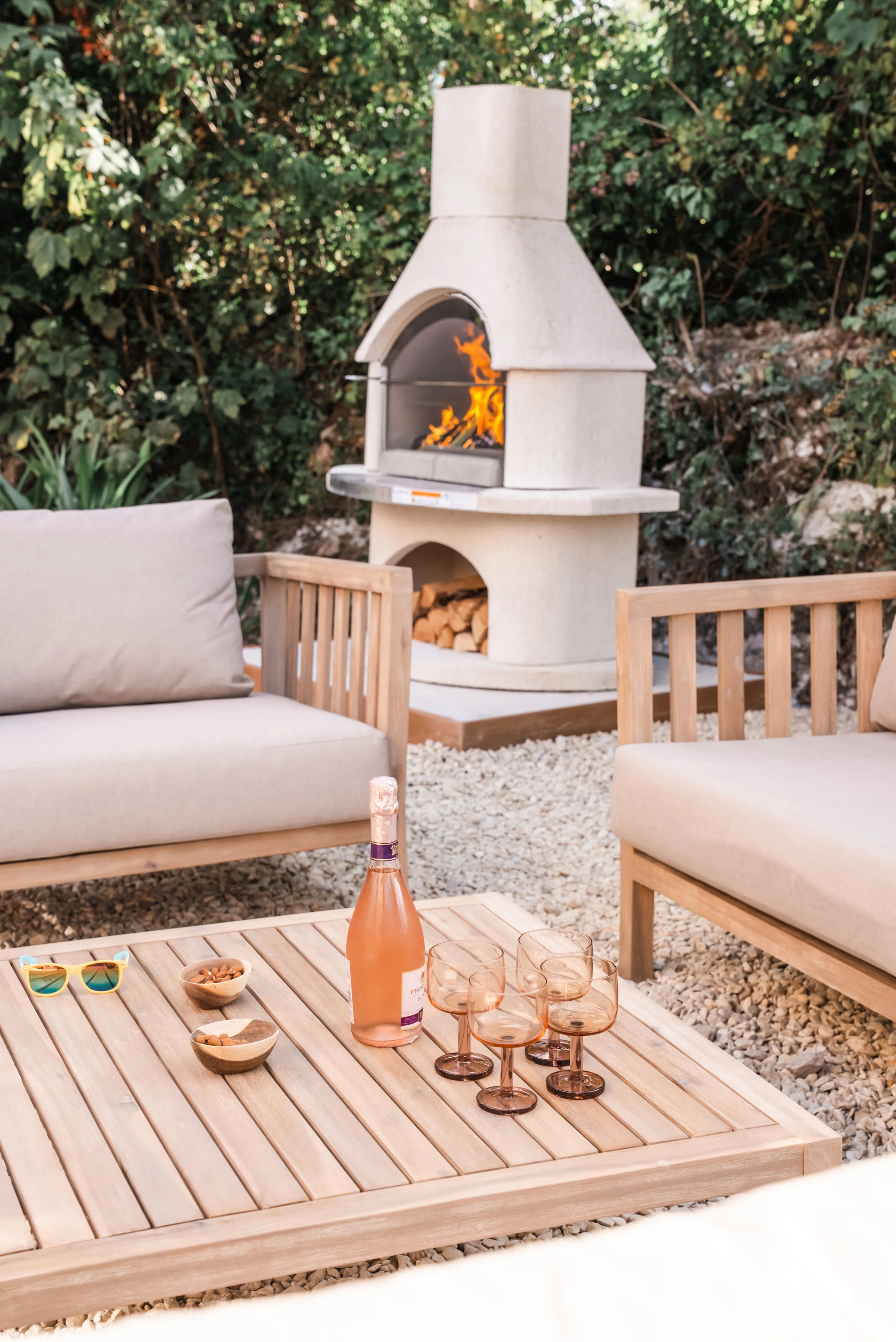 A relaxed outdoor seating area arranged around a sculptural wood-fired oven, creating a natural focal point for social moments. Soft upholstered furniture, warm timber surfaces, and loose gravel underfoot combine to form an inviting setting that enco