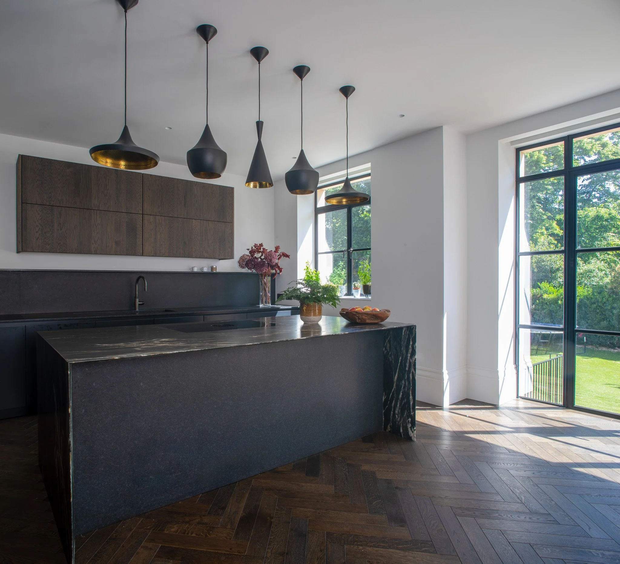 This contemporary kitchen centres around a sculptural stone island, paired with dark timber cabinetry and soft matte finishes. A series of pendant lights brings rhythm and warmth to the space, while generous glazing floods the room with daylight and 
